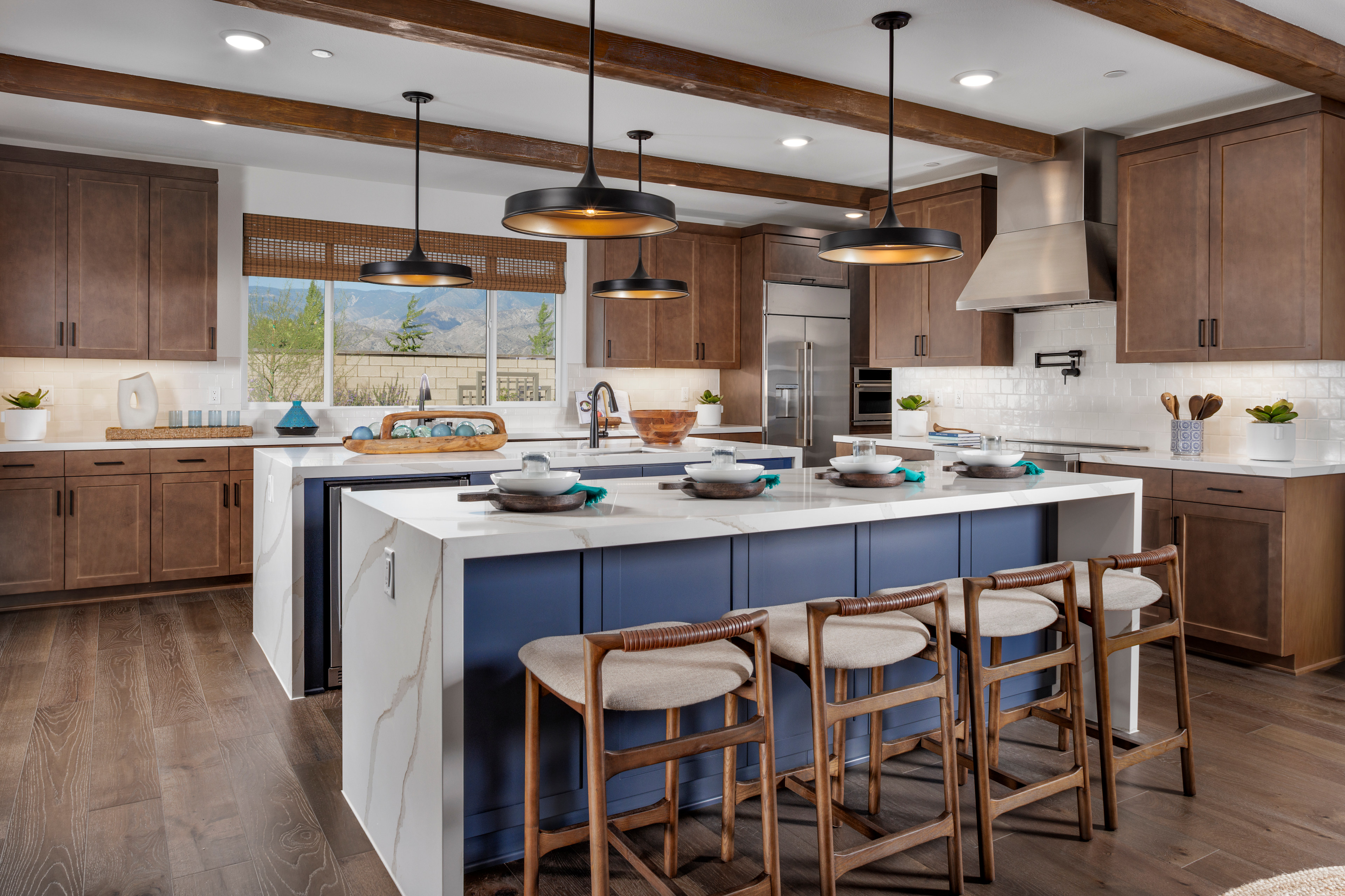 A modern and spacious kitchen with a large central island, wooden cabinets, pendant lights, and a view of the outdoors through a window.