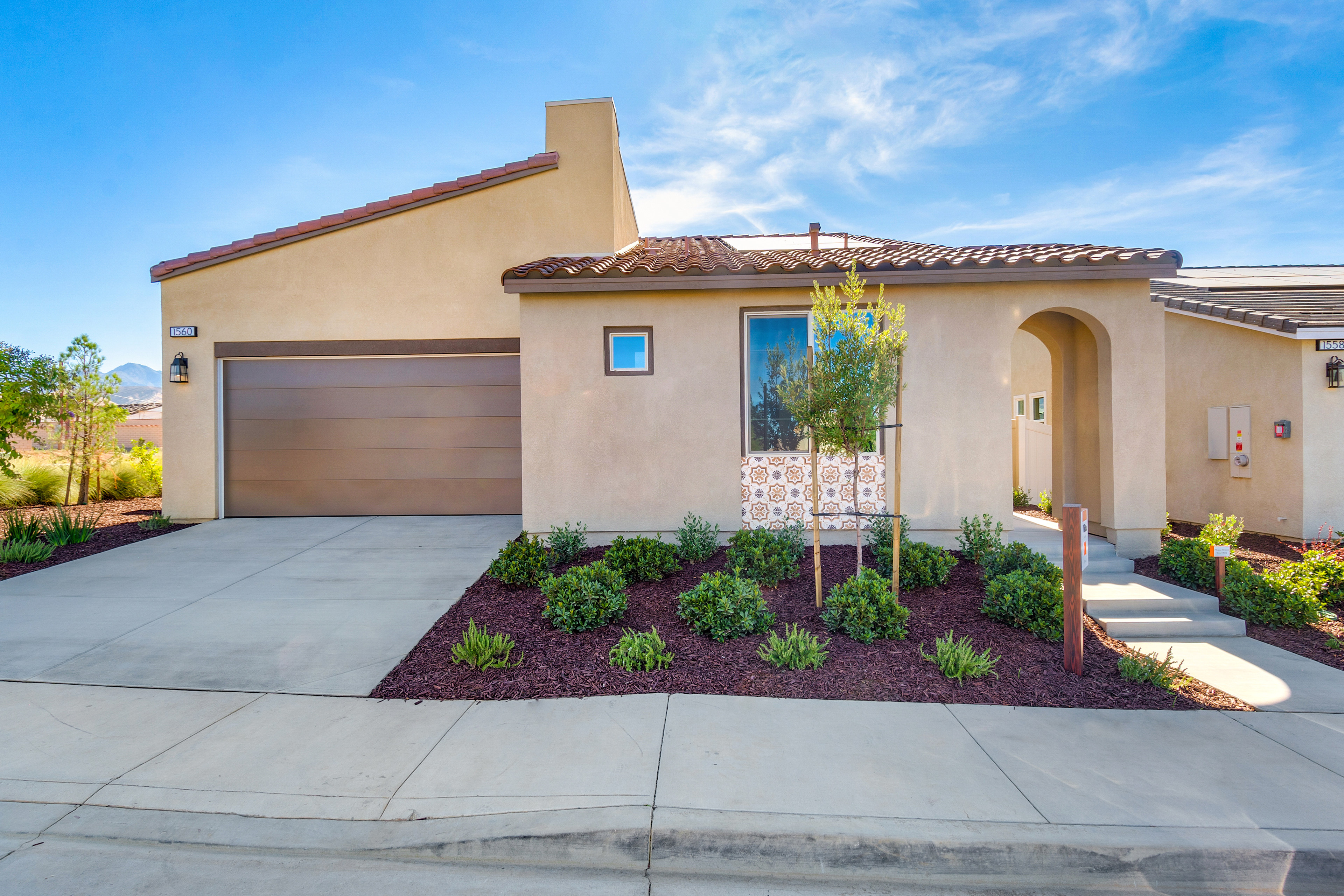 A modern, single-story house with a tiled roof, a garage door, and a well-landscaped front yard featuring various plants and a concrete walkway leading to the entrance.