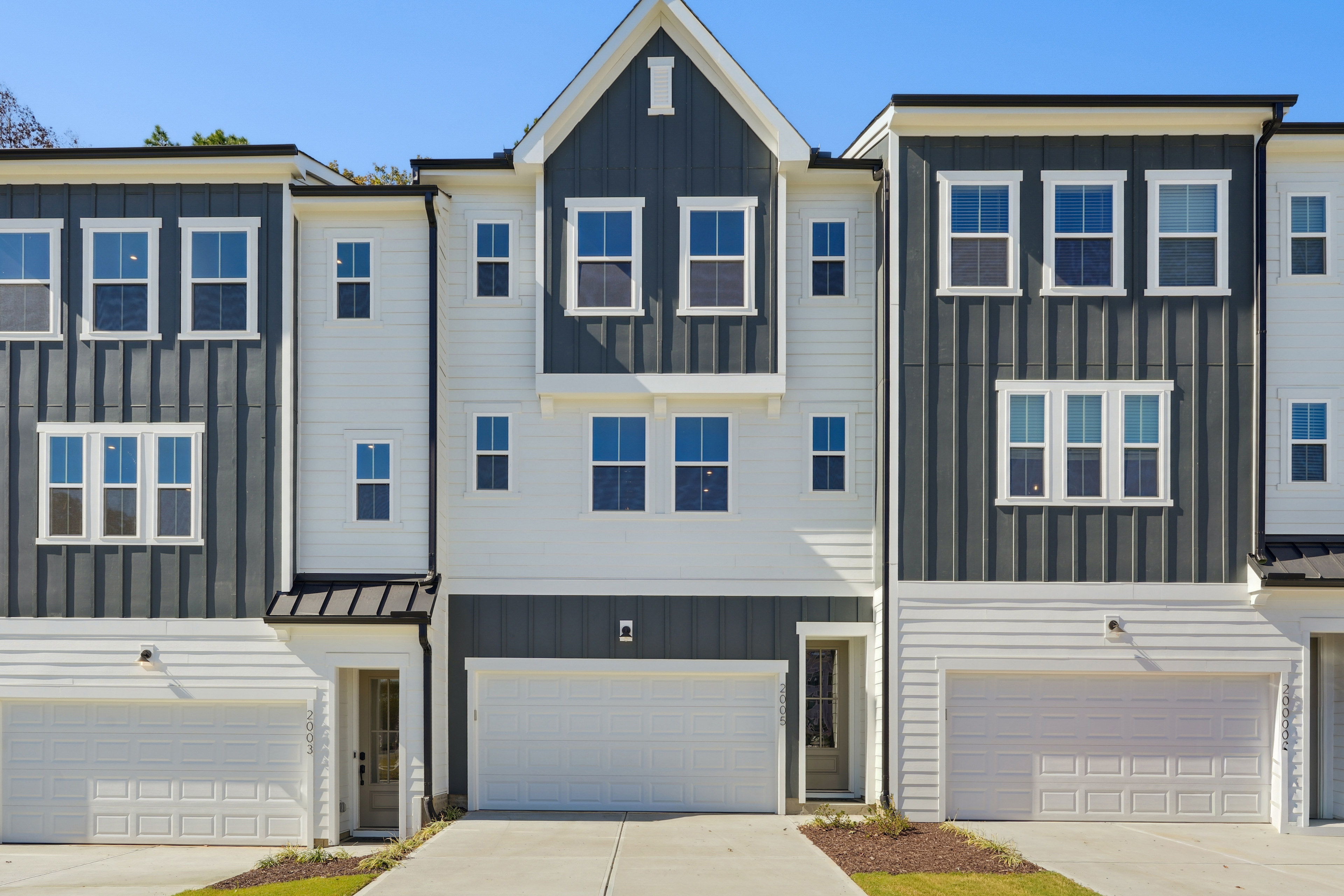 A modern, multi-story townhouse with a gray and white exterior, featuring a prominent gable roof and multiple garage doors in the foreground, set against a backdrop of a clear blue sky.