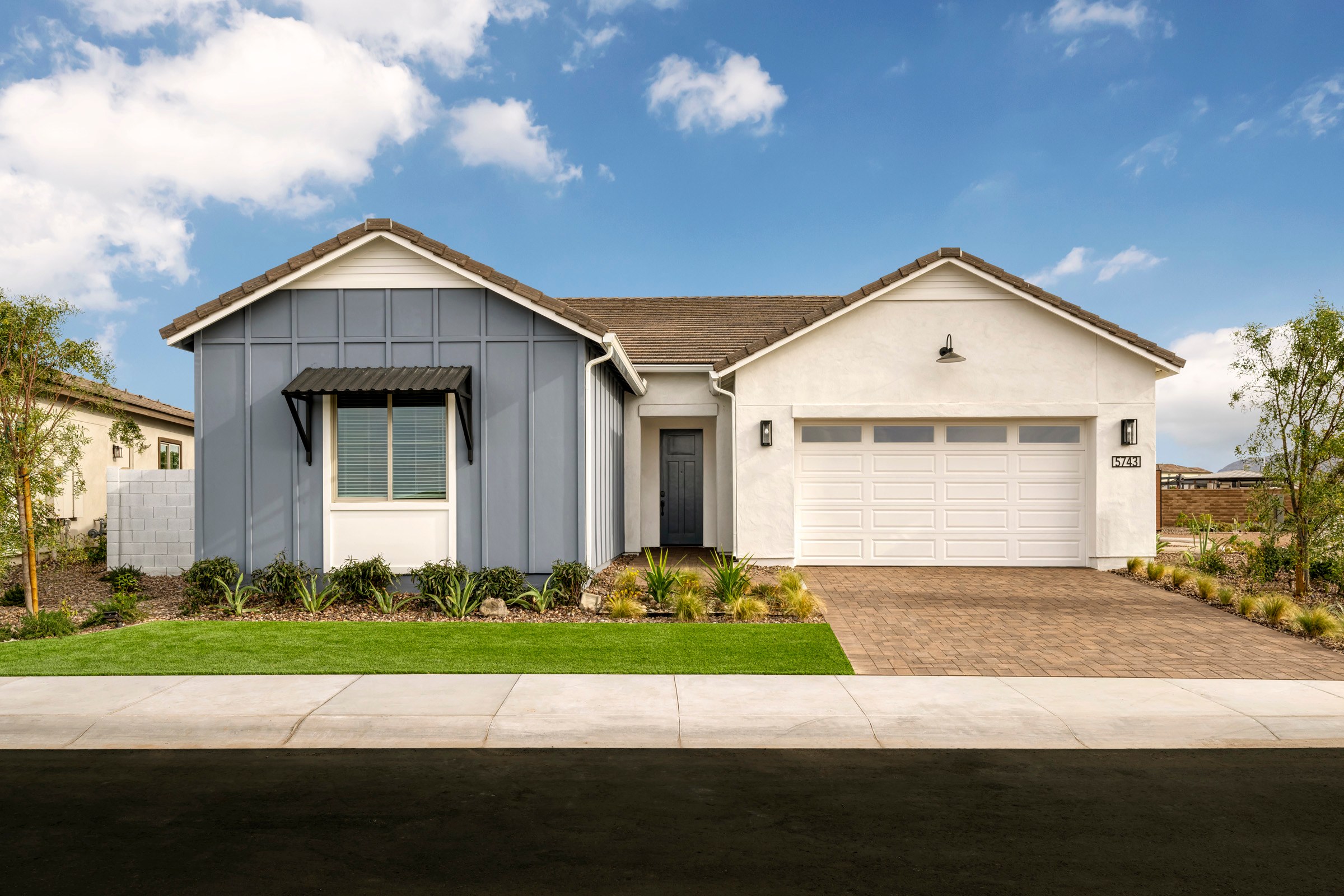 A modern, two-story house with a gray and white exterior, a garage, and a well-manicured lawn in the foreground, set against a blue sky with fluffy white clouds.