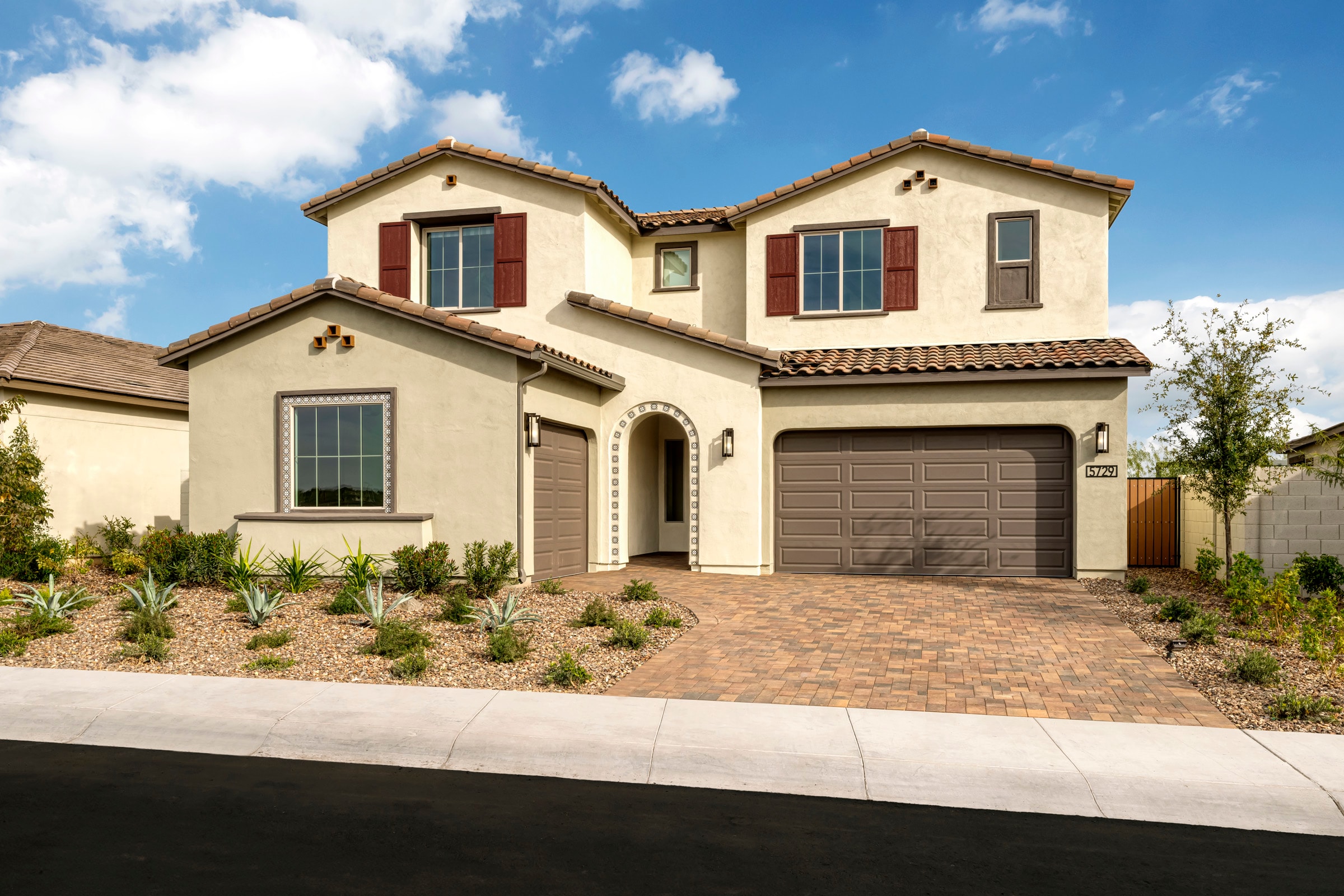 A two-story Mediterranean-style house with a red tile roof, stucco exterior, and a garage door stands in a landscaped yard with shrubs and trees against a blue sky with fluffy white clouds.