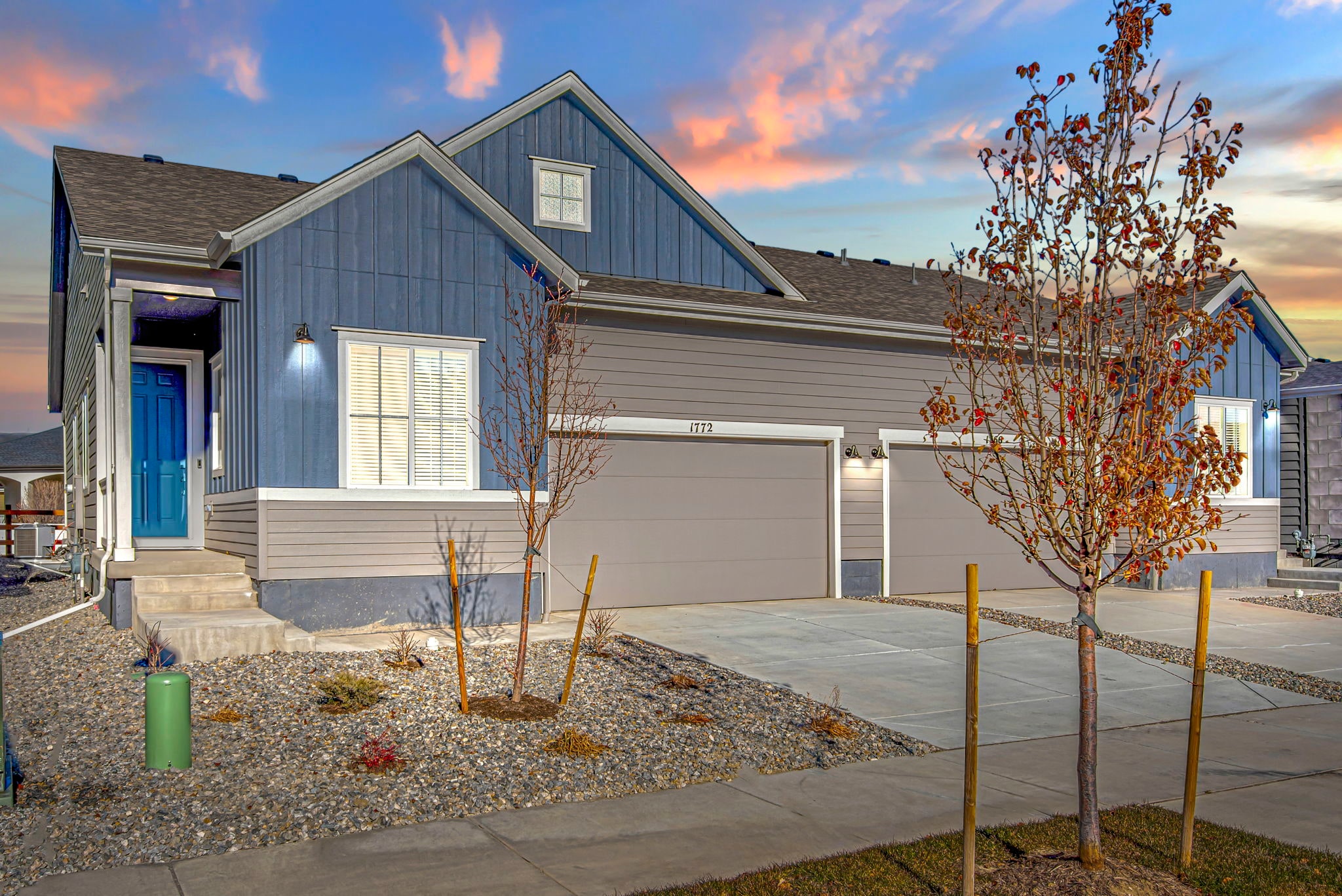 A modern two-story house with a gray exterior, a blue front door, and a colorful autumn tree in the foreground, set against a vibrant sunset sky.