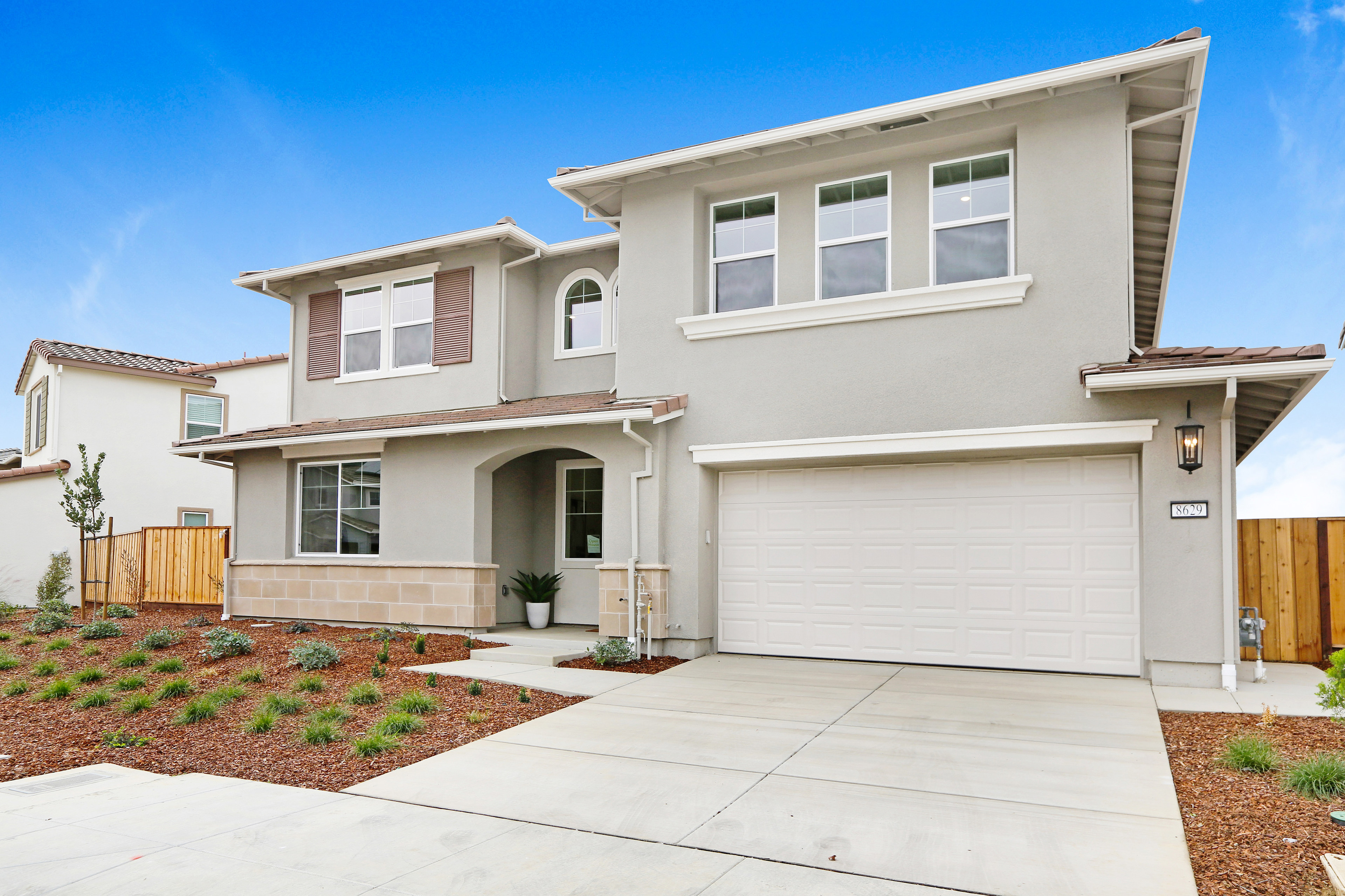 A two-story beige house with a garage door, surrounded by a landscaped yard and a clear blue sky in the background.