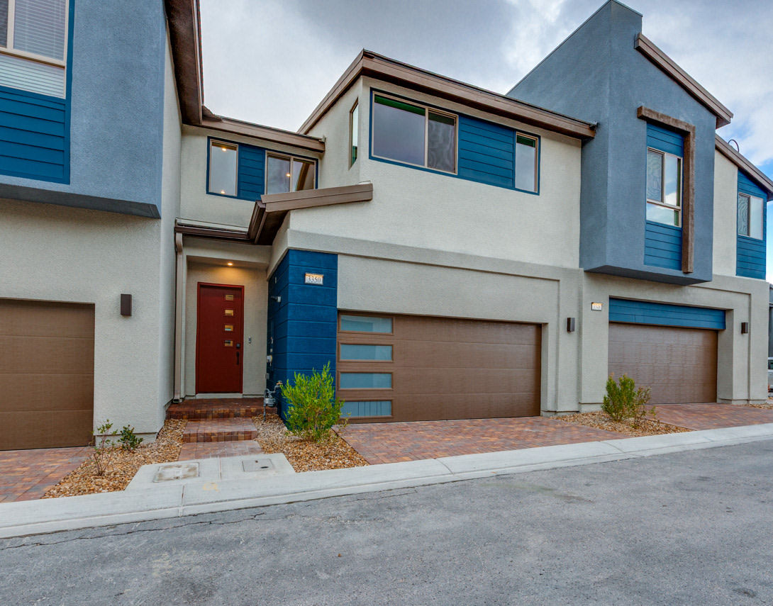 A modern, multi-story townhouse with a garage and a landscaped front yard, featuring a mix of neutral and blue tones in its exterior design.