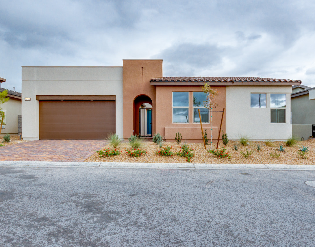 A modern, single-story house with a garage, featuring a stucco exterior, a tiled roof, and a landscaped front yard against a cloudy sky backdrop.