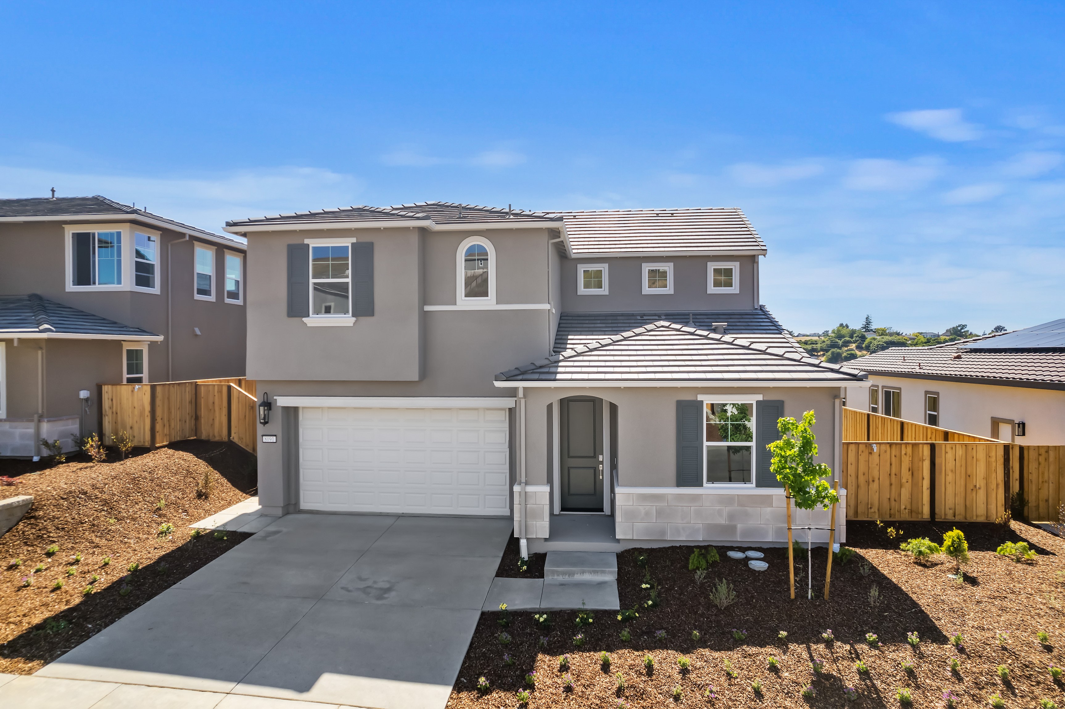 A newly constructed two-story residential home with a garage, surrounded by a freshly landscaped yard and neighboring houses in a suburban setting.