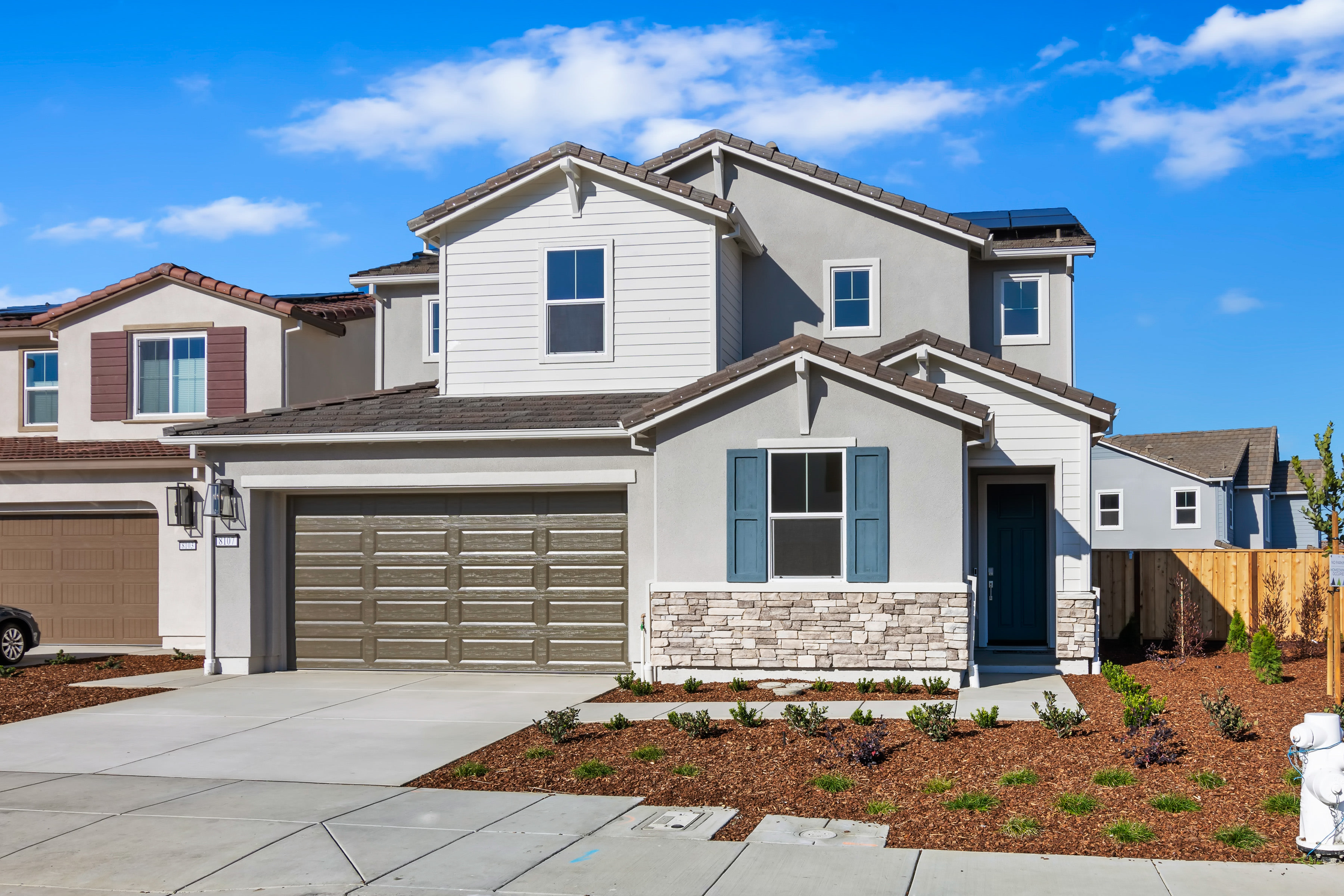 A two-story residential house with a garage, surrounded by landscaped yards and a clear blue sky with scattered clouds.