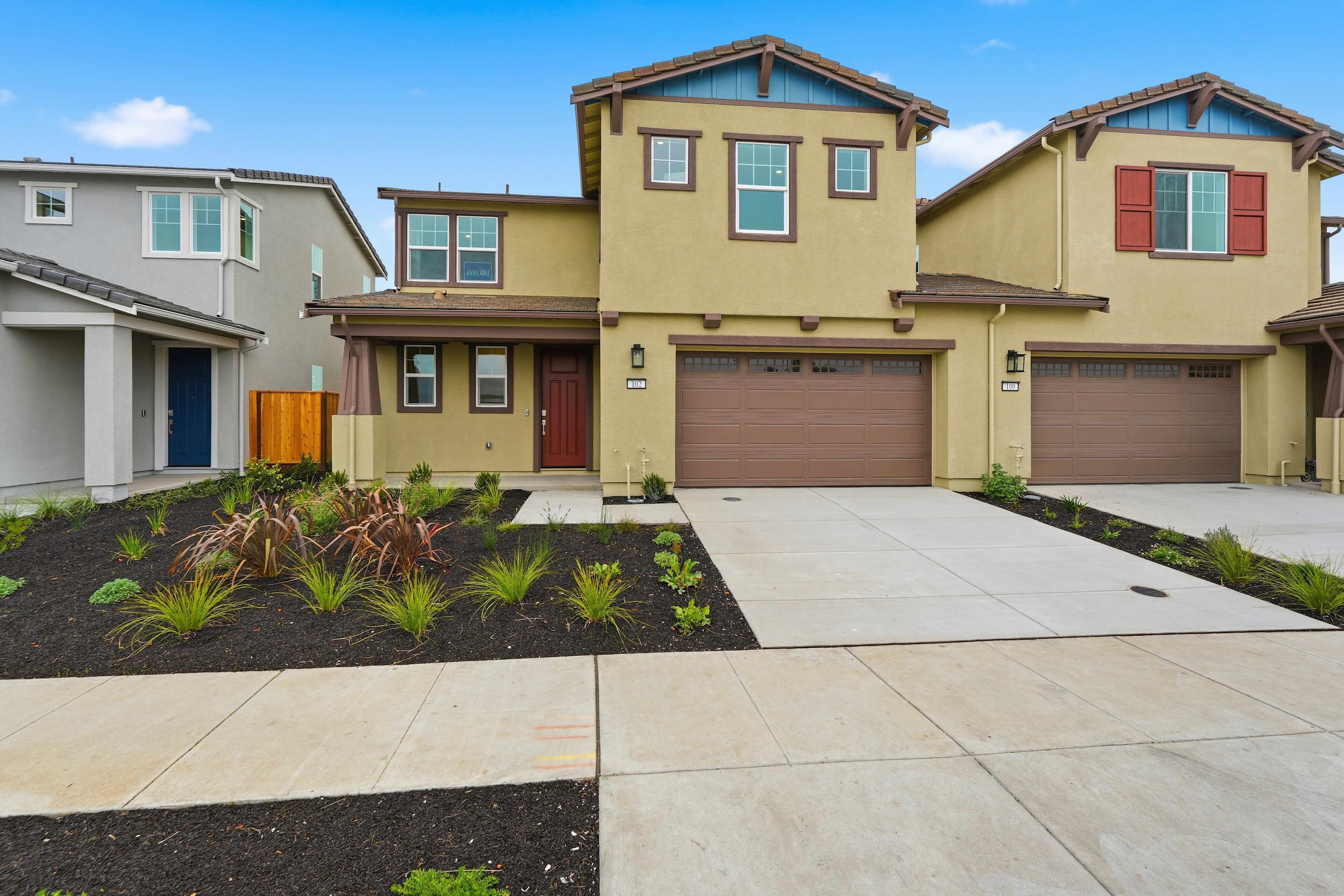 A modern, multi-story residential building with a garage and a well-landscaped front yard, set against a clear blue sky.
