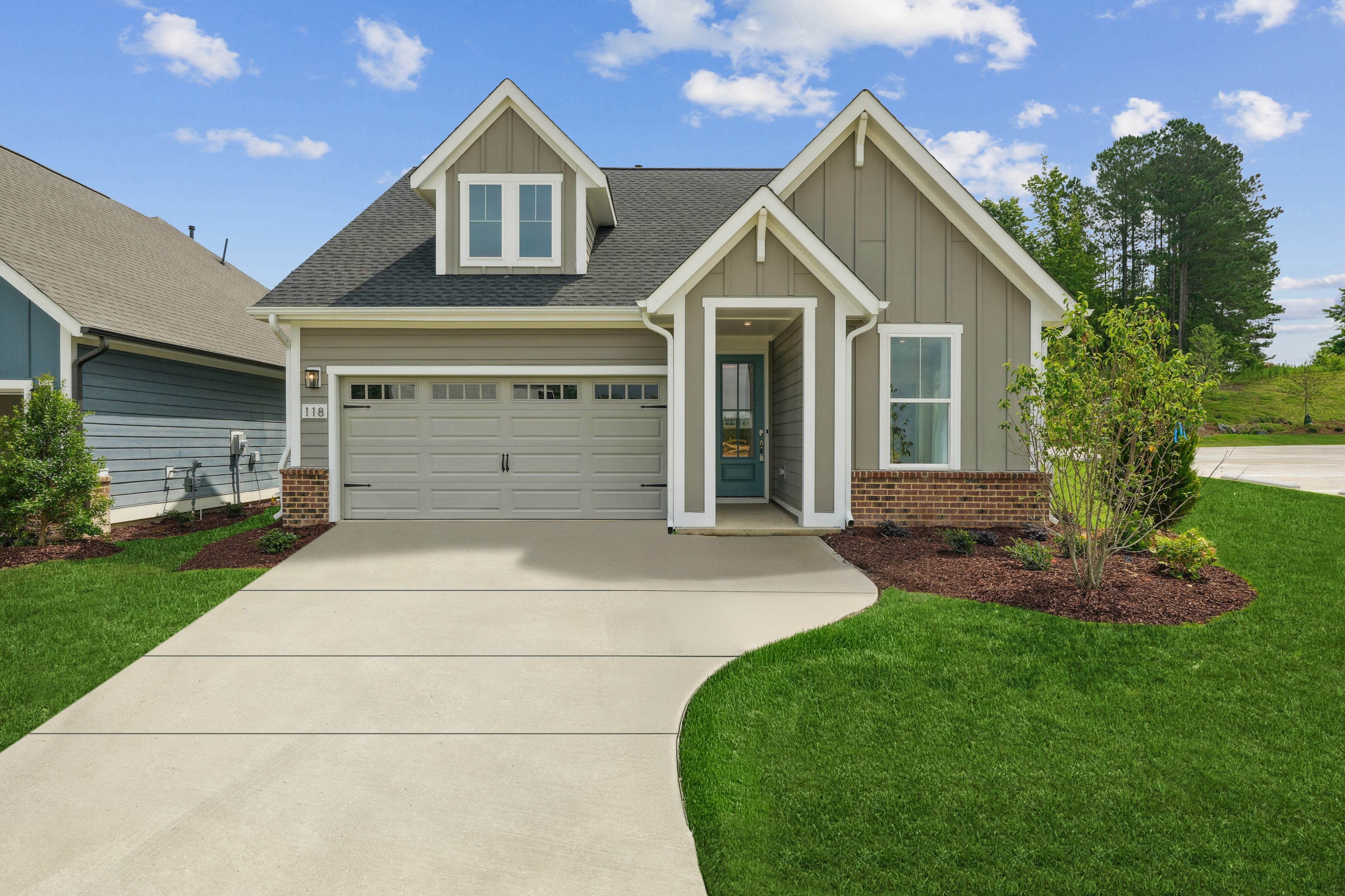 A well-manicured lawn leads to a two-story house with a gray exterior, a garage, and a lush green landscape in the background.