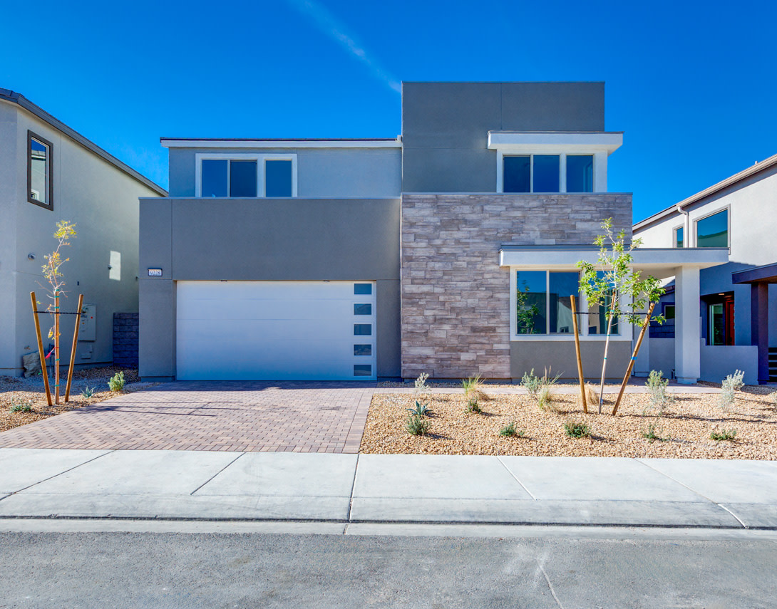 A modern, two-story residential building with a gray exterior, large windows, and a paved driveway in the foreground, set against a clear blue sky.
