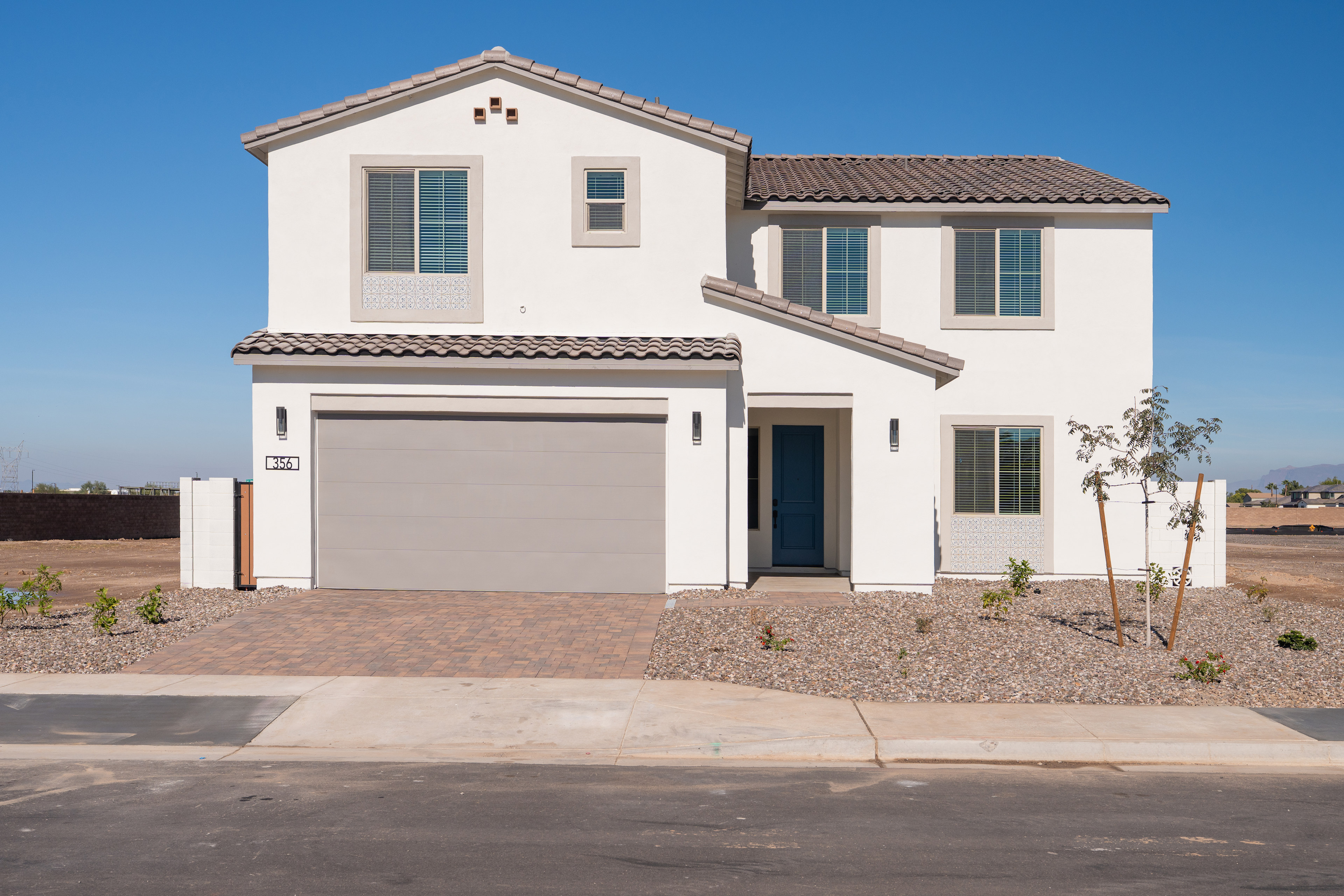 A two-story white stucco house with a tiled roof, a garage door, and a paved driveway, set against a clear blue sky in the background.