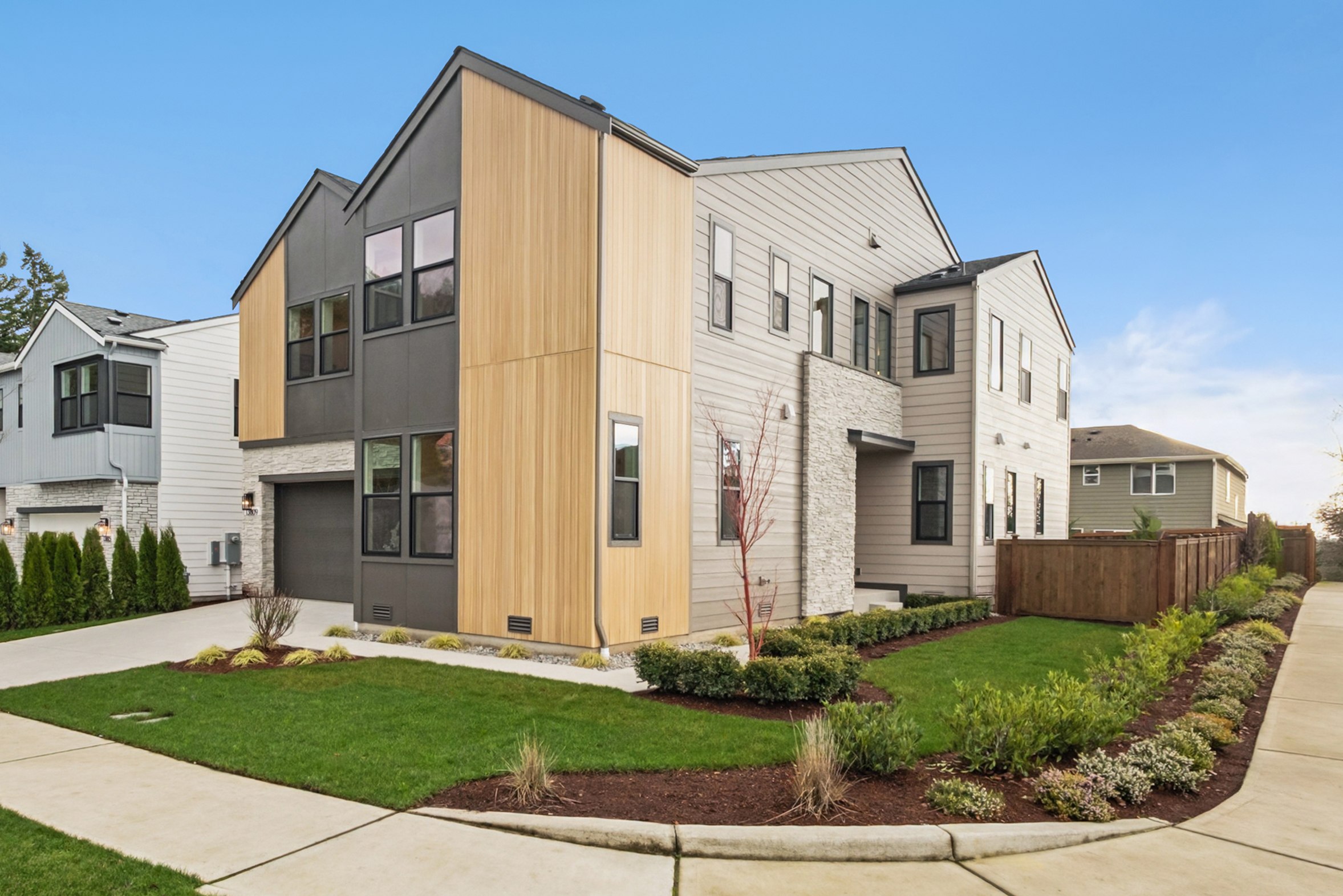 A modern, multi-story residential building with a well-landscaped front yard, surrounded by other similar structures in a suburban neighborhood setting.