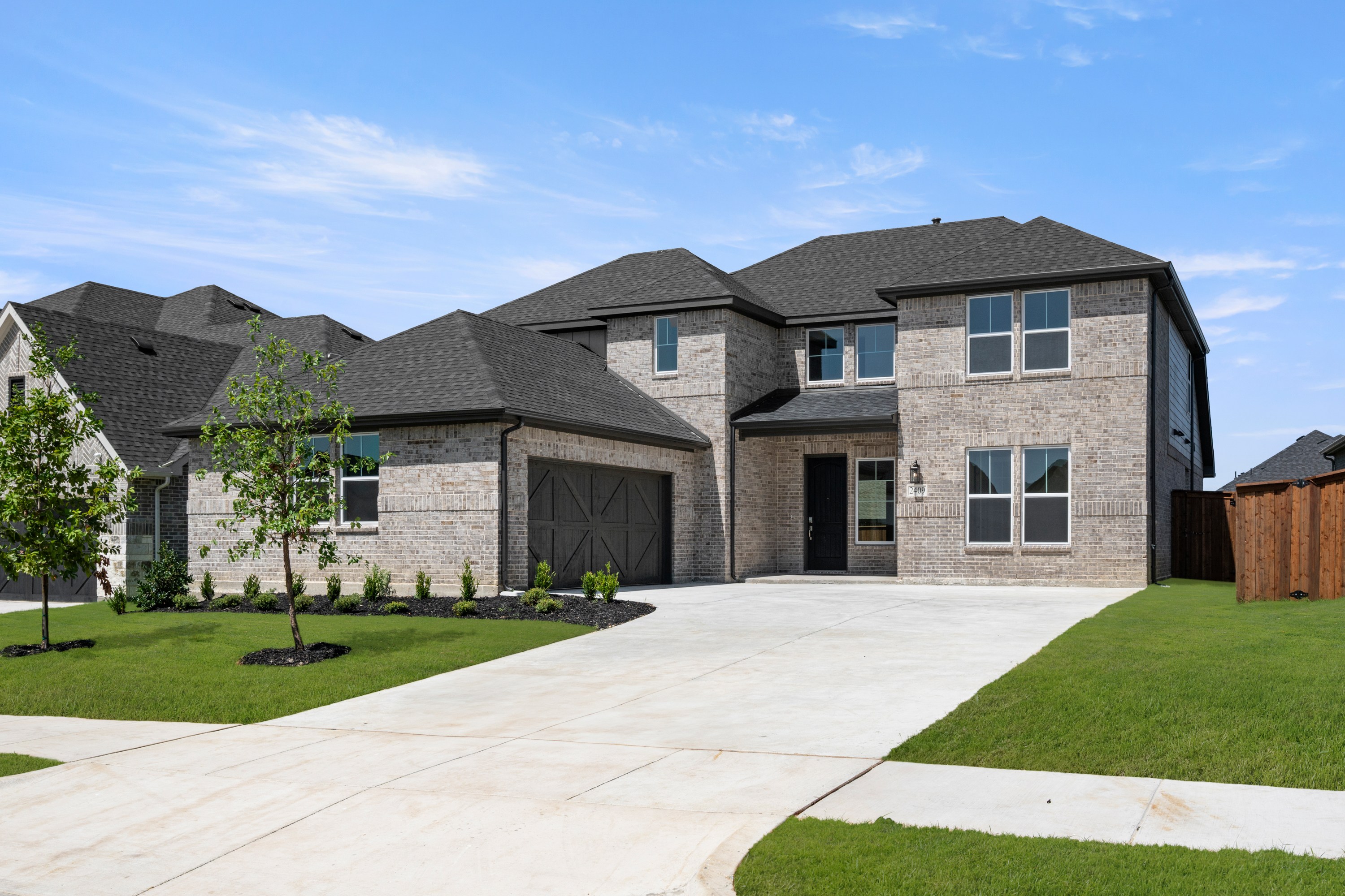 A large, two-story brick house with a well-manicured lawn and a paved driveway leading to the front entrance, set against a clear blue sky with scattered clouds.