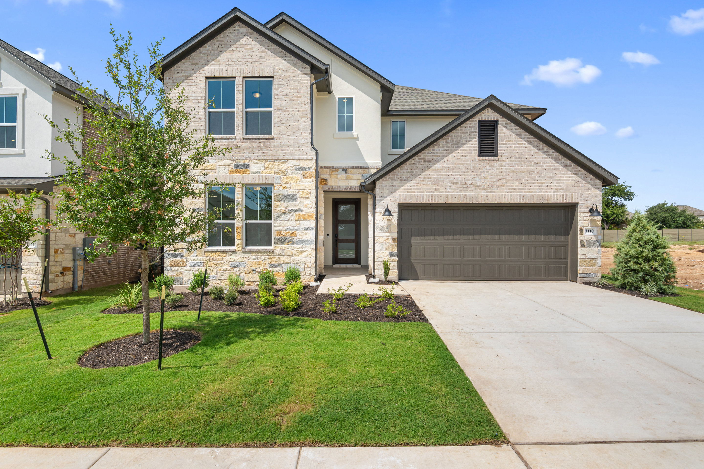A two-story residential house with a stone exterior, a garage, and a well-manicured lawn in the foreground, set against a clear blue sky with some clouds.