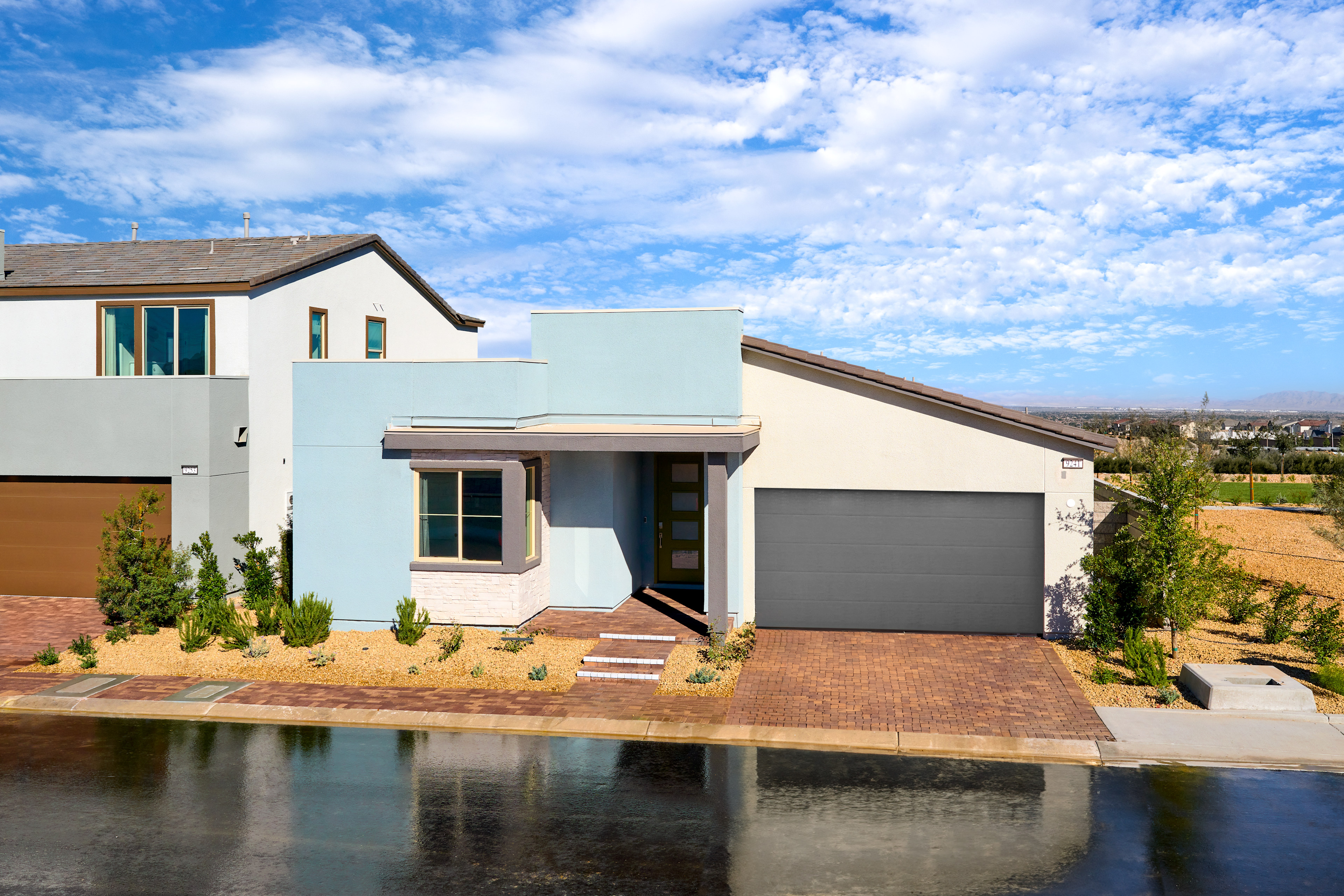 A modern, two-story house with a garage and a small pond in the foreground, set against a backdrop of a blue sky with fluffy white clouds.