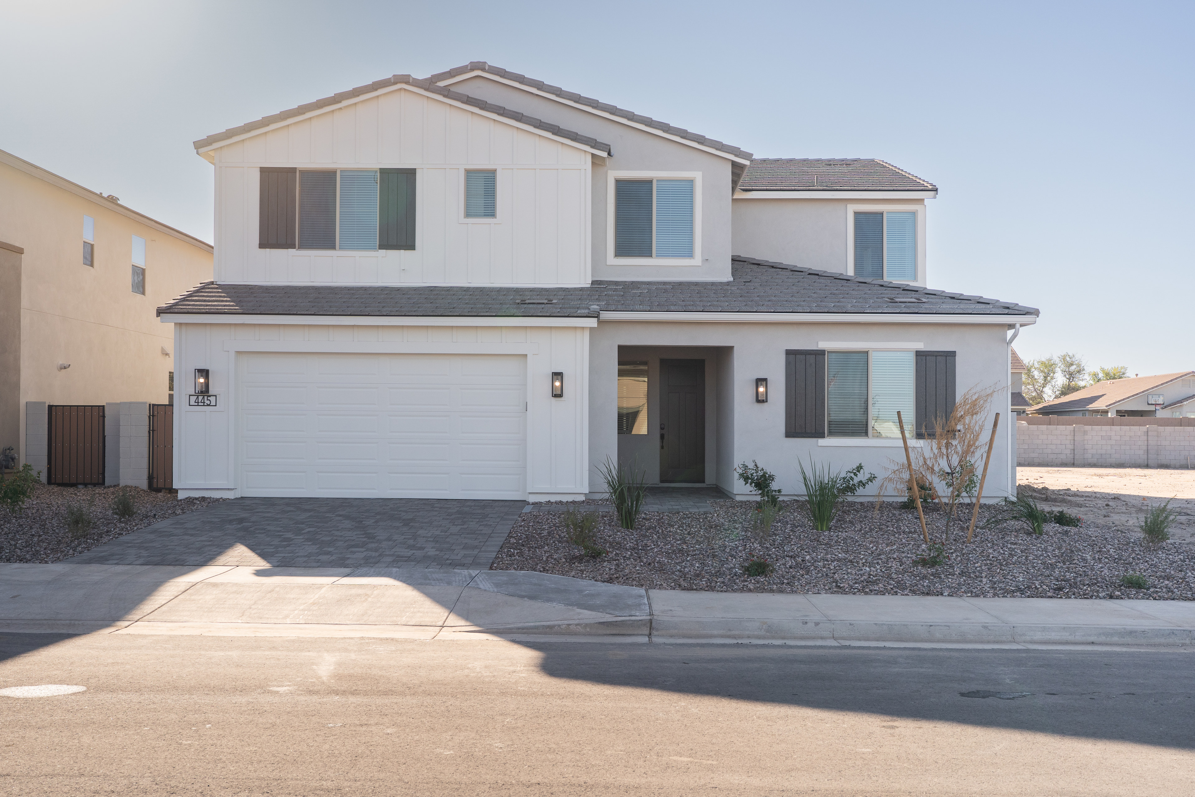 A two-story residential house with a garage, surrounded by a paved driveway and landscaped yard, set against a clear blue sky.