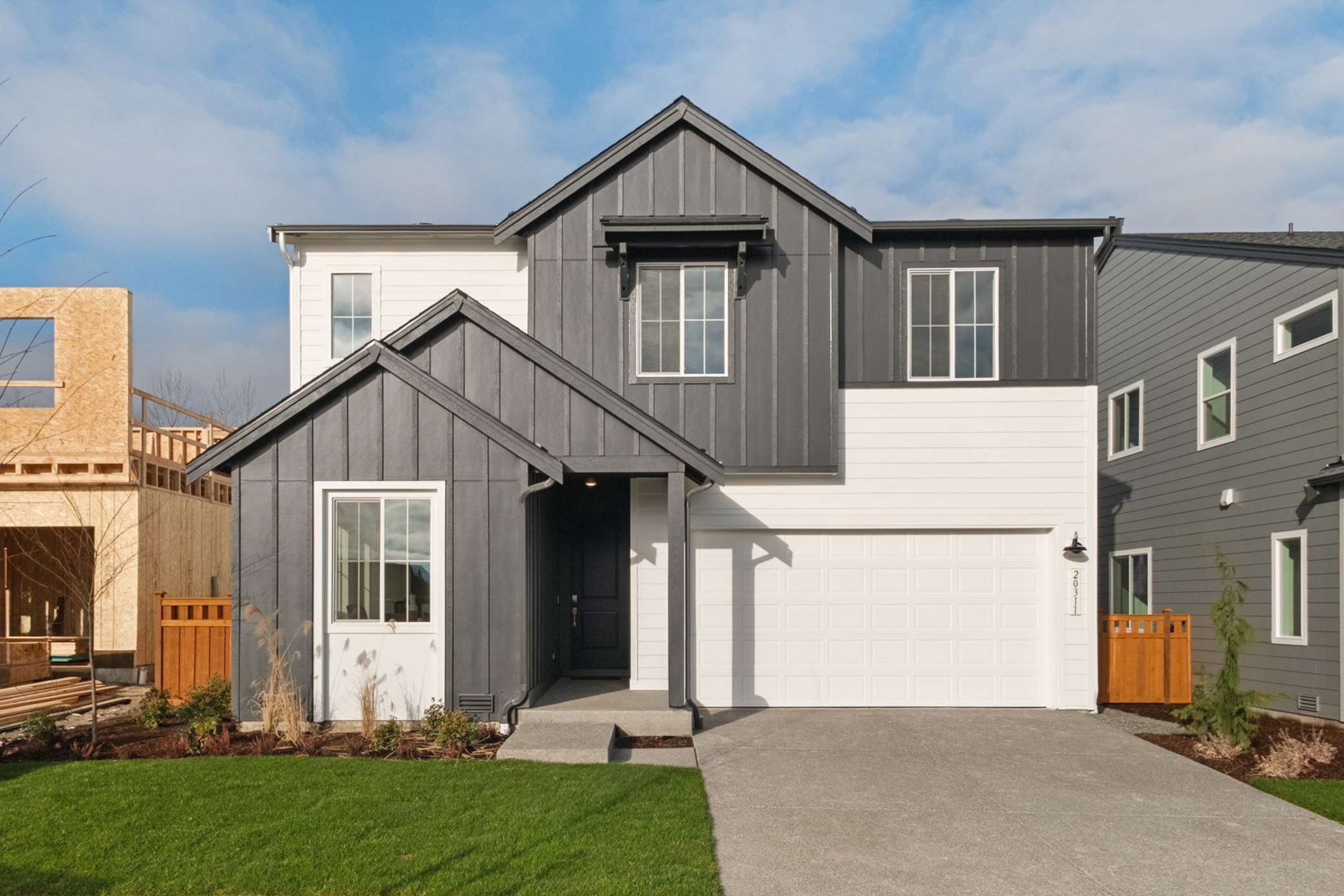 A modern two-story house with a gray exterior, a white garage door, and a well-manicured lawn in the foreground, set against a blue sky with scattered clouds.
