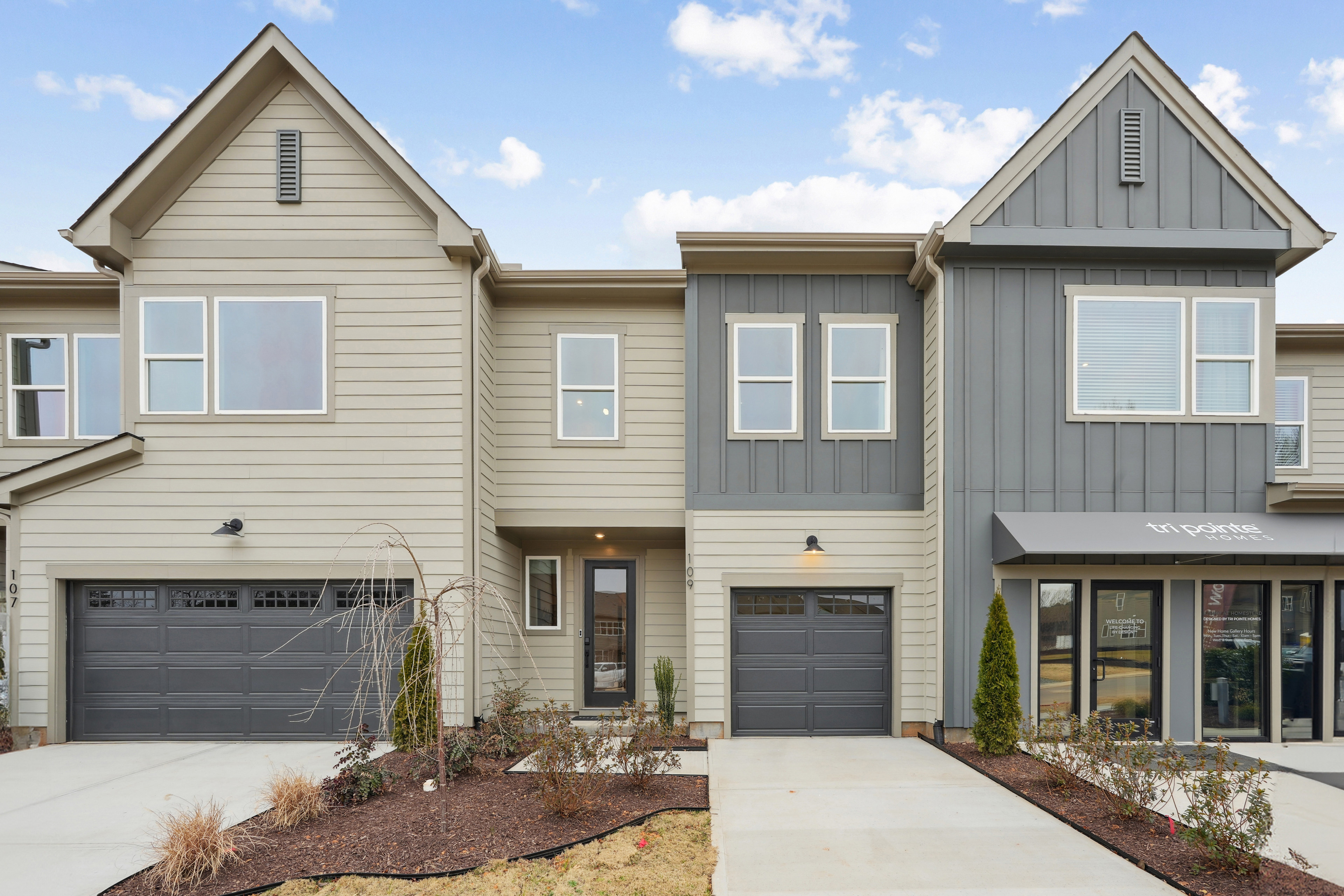 The image depicts a modern townhouse complex with two-story buildings featuring gabled roofs, siding in neutral tones, and attached garages. The foreground includes a paved walkway and landscaping elements such as plants and shrubs.