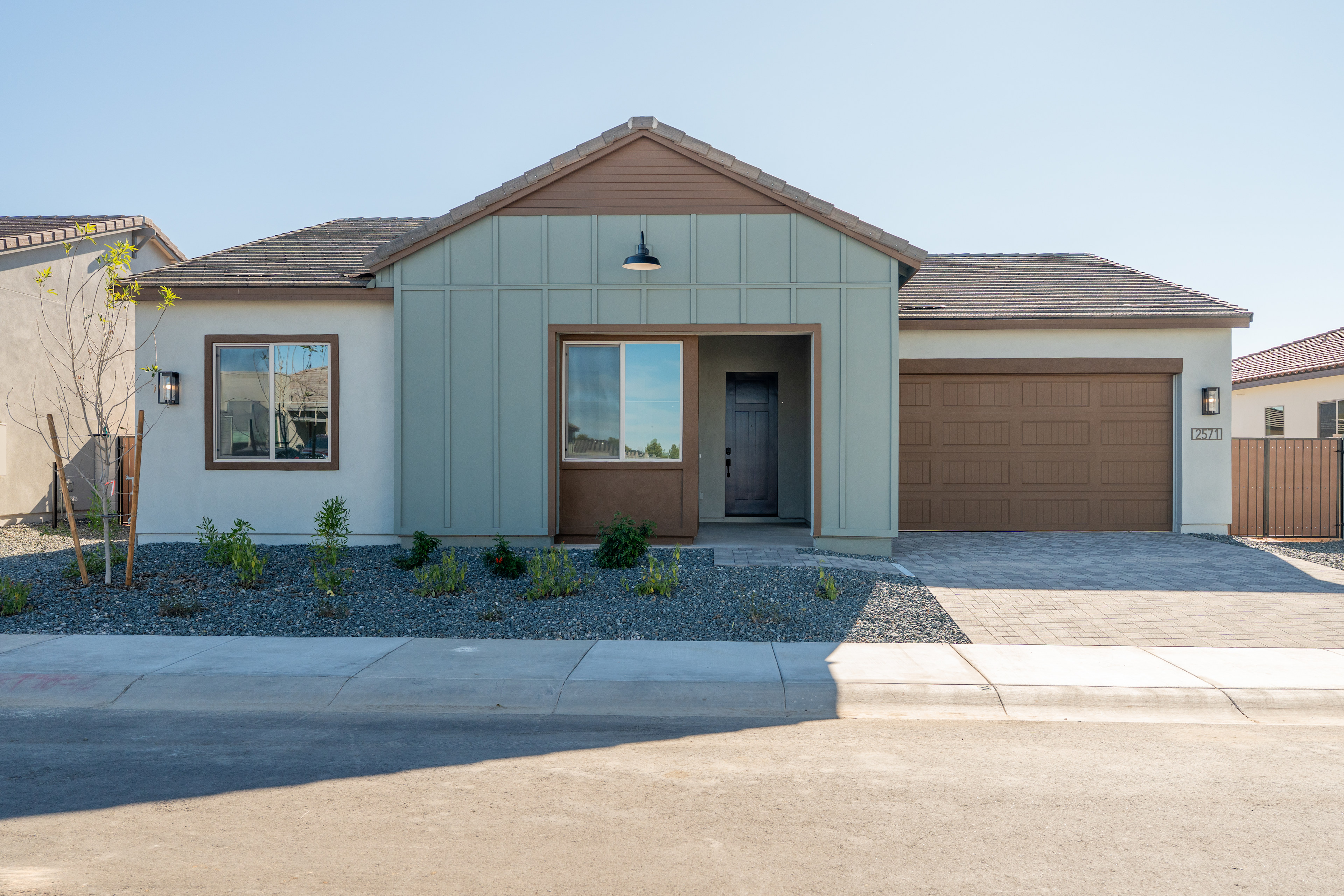 A modern, single-story house with a gabled roof, siding in neutral tones, and a paved driveway leading to a garage door in the foreground. The house is surrounded by landscaping, including shrubs and a paved walkway.