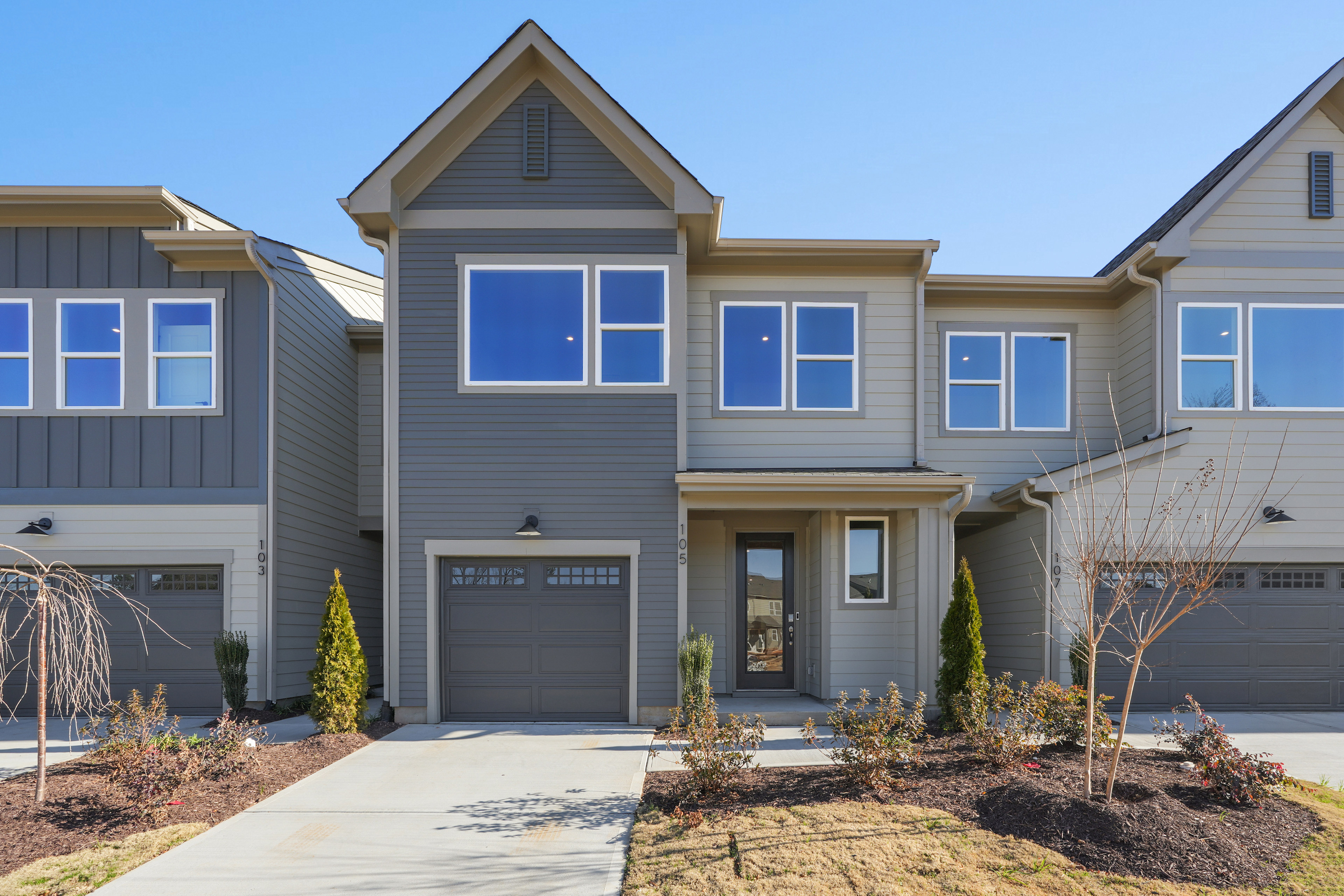 A modern two-story townhouse with a gray exterior, a gabled roof, and a well-landscaped front yard with shrubs and trees.