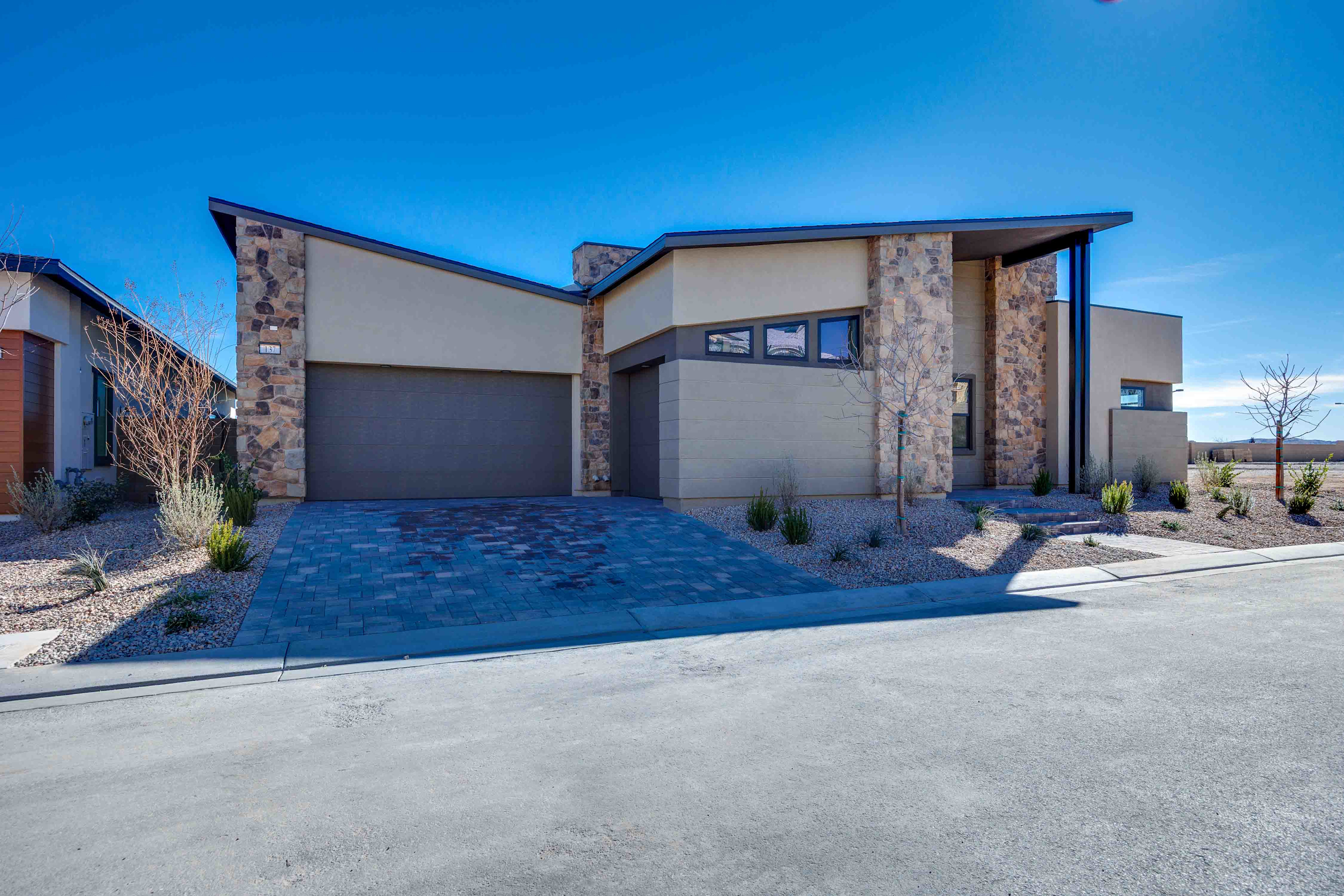 A modern, two-story house with a stone exterior, a garage, and a paved driveway in the foreground, set against a clear blue sky in the background.