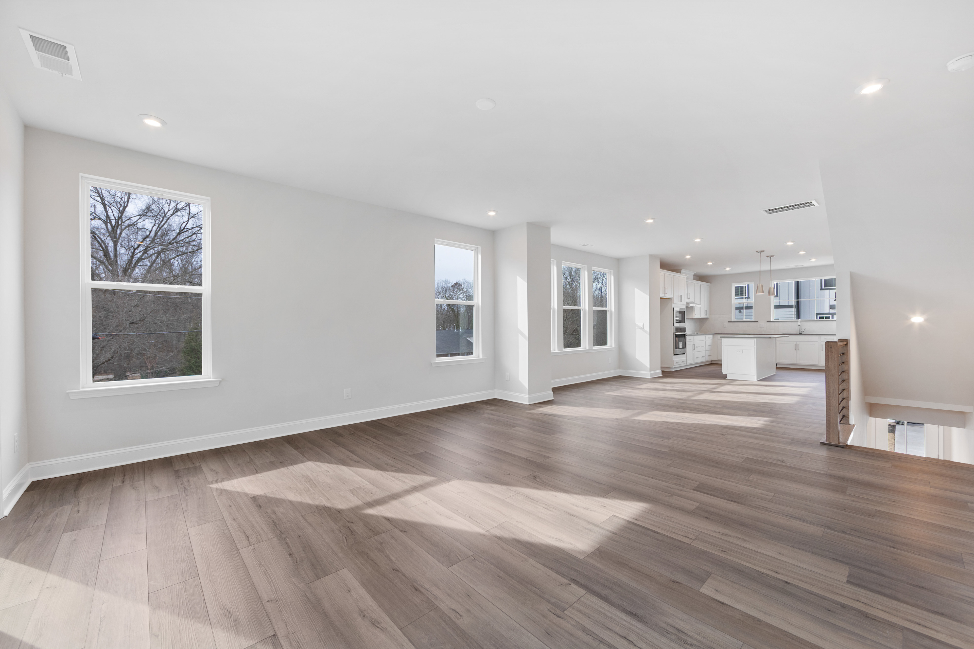 A spacious, open-concept living area with hardwood floors, large windows, and a kitchen visible in the background.