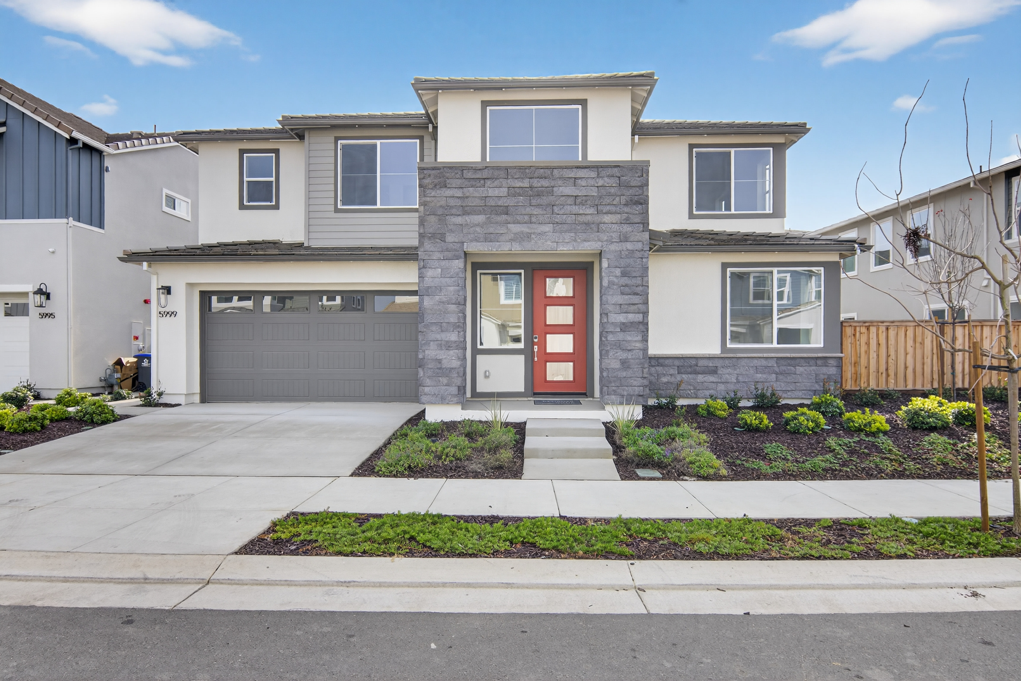 A modern two-story residential house with a gray exterior, a red front door, and a well-landscaped front yard with plants and a paved driveway, set against a clear blue sky with fluffy white clouds.