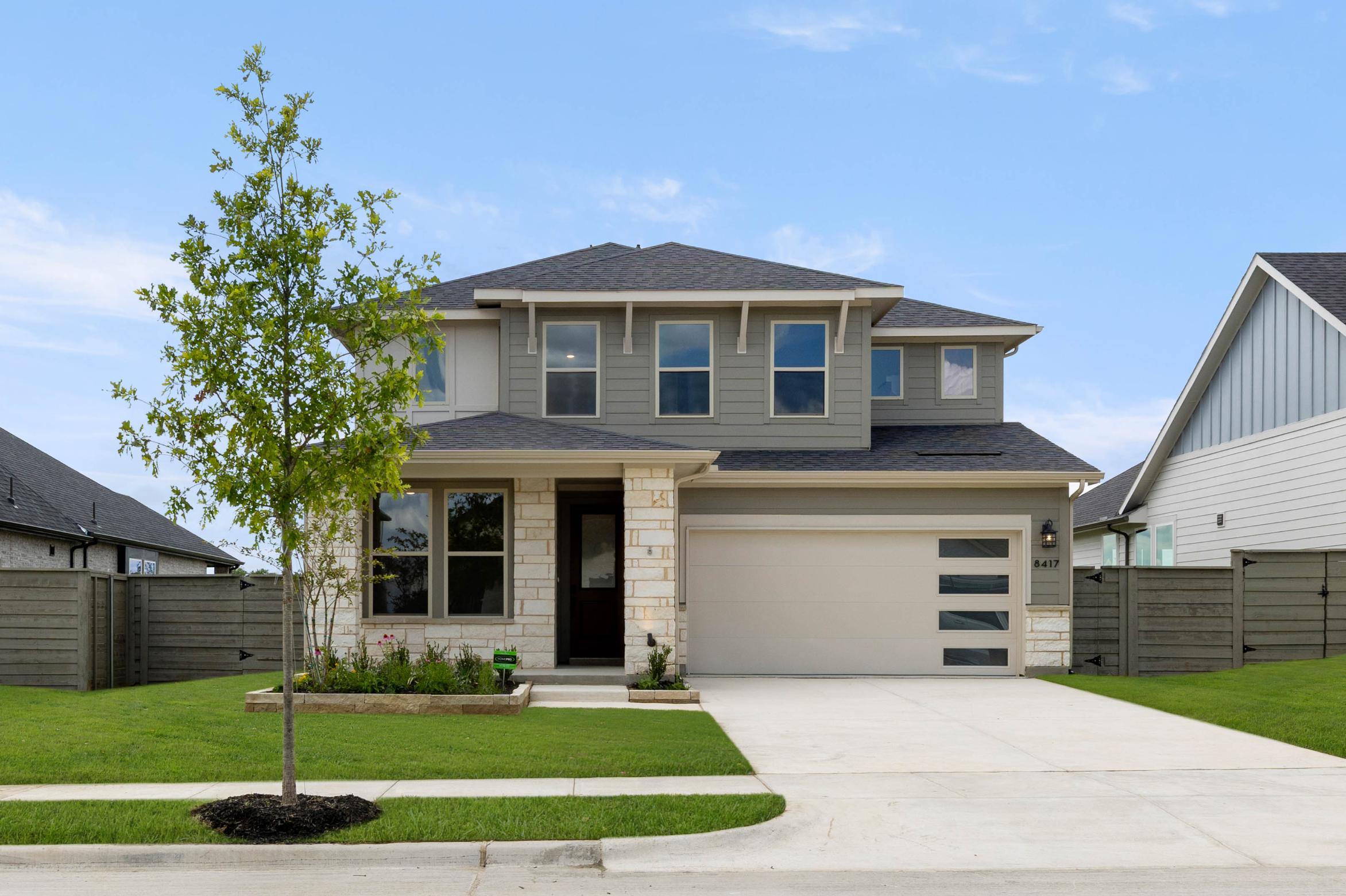 A two-story residential house with a well-manicured lawn, surrounded by trees and a wooden fence in the background.