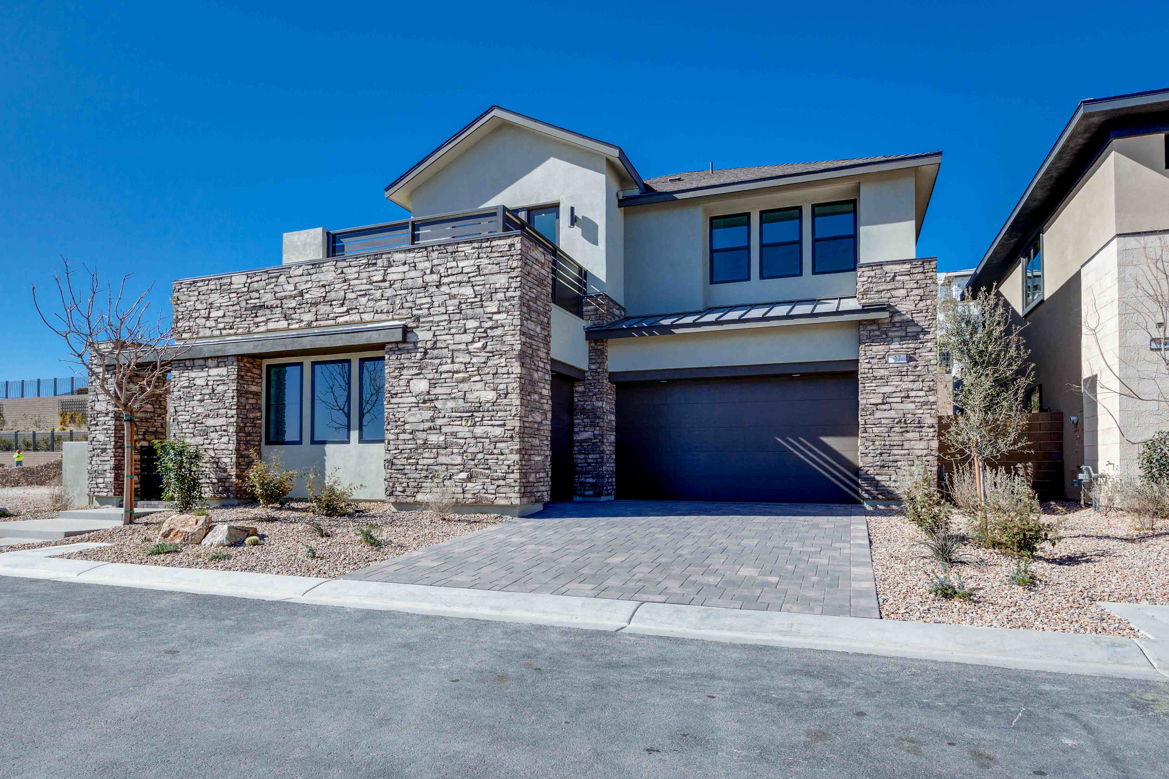 A modern two-story house with a stone exterior, a garage door, and a paved driveway in the foreground, set against a clear blue sky in the background.