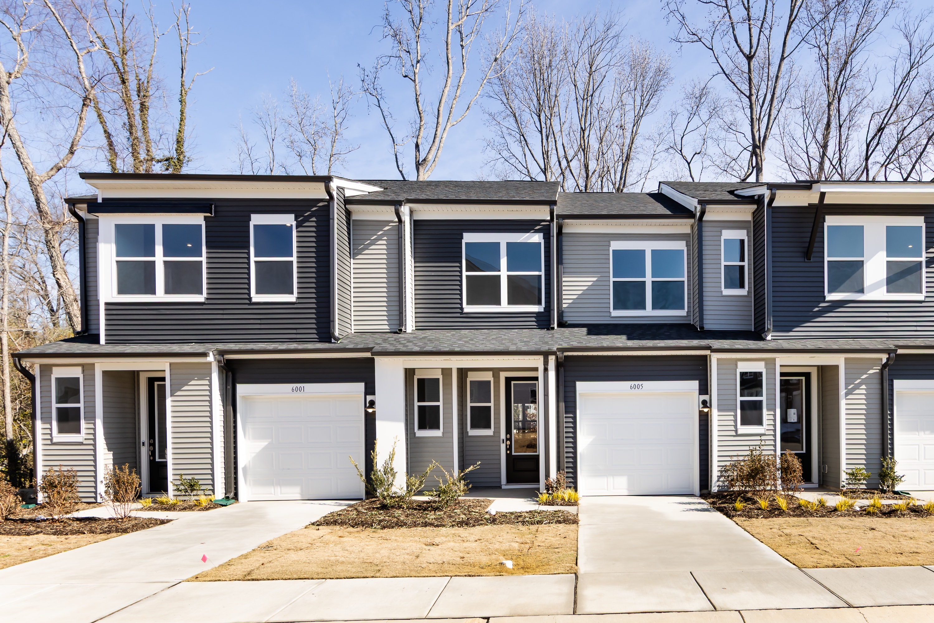 The image depicts a row of modern townhouses with gray siding and white trim, set against a backdrop of bare trees and a clear blue sky.