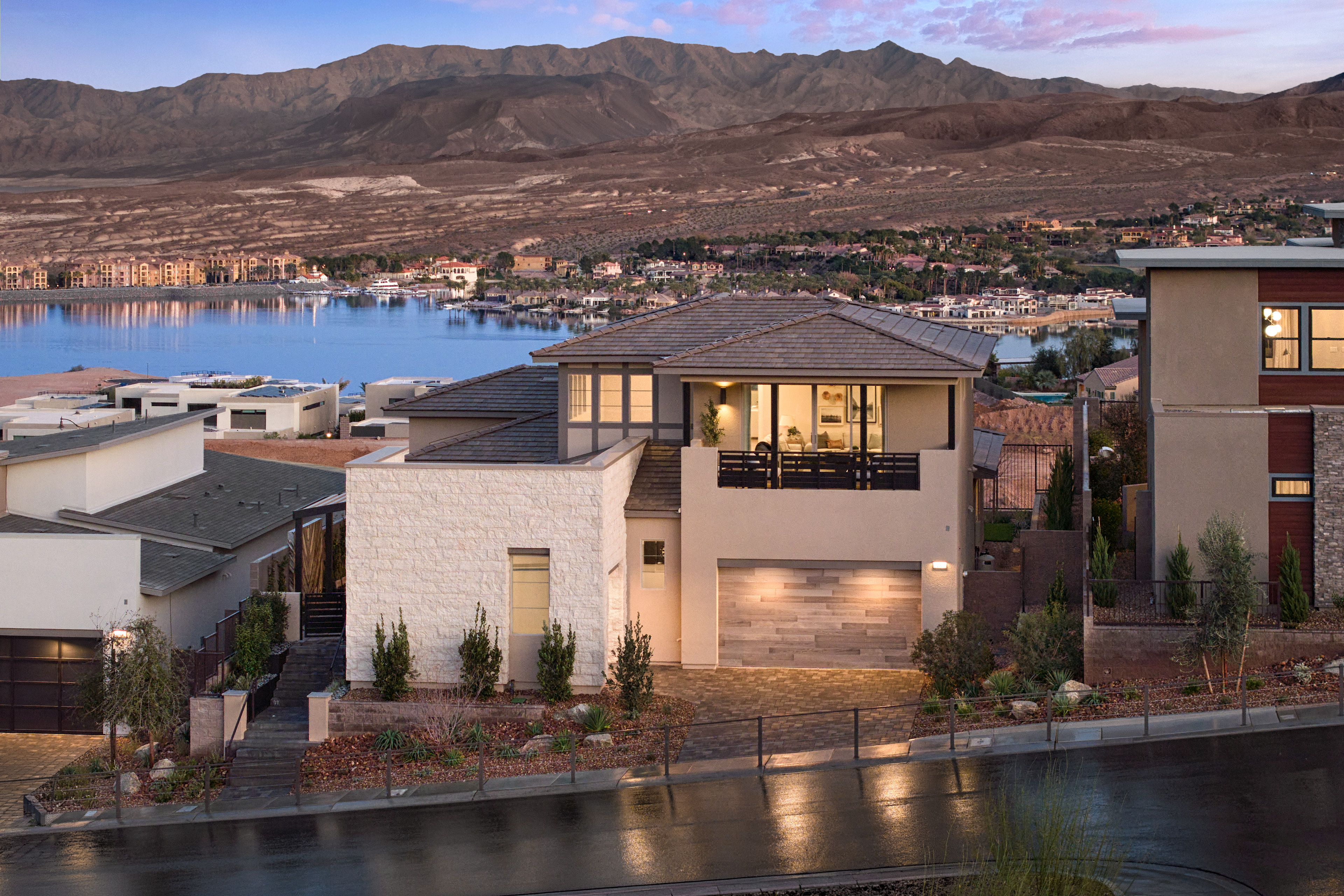 A modern, multi-story house with a balcony overlooking a scenic lake surrounded by rugged mountains in the background.