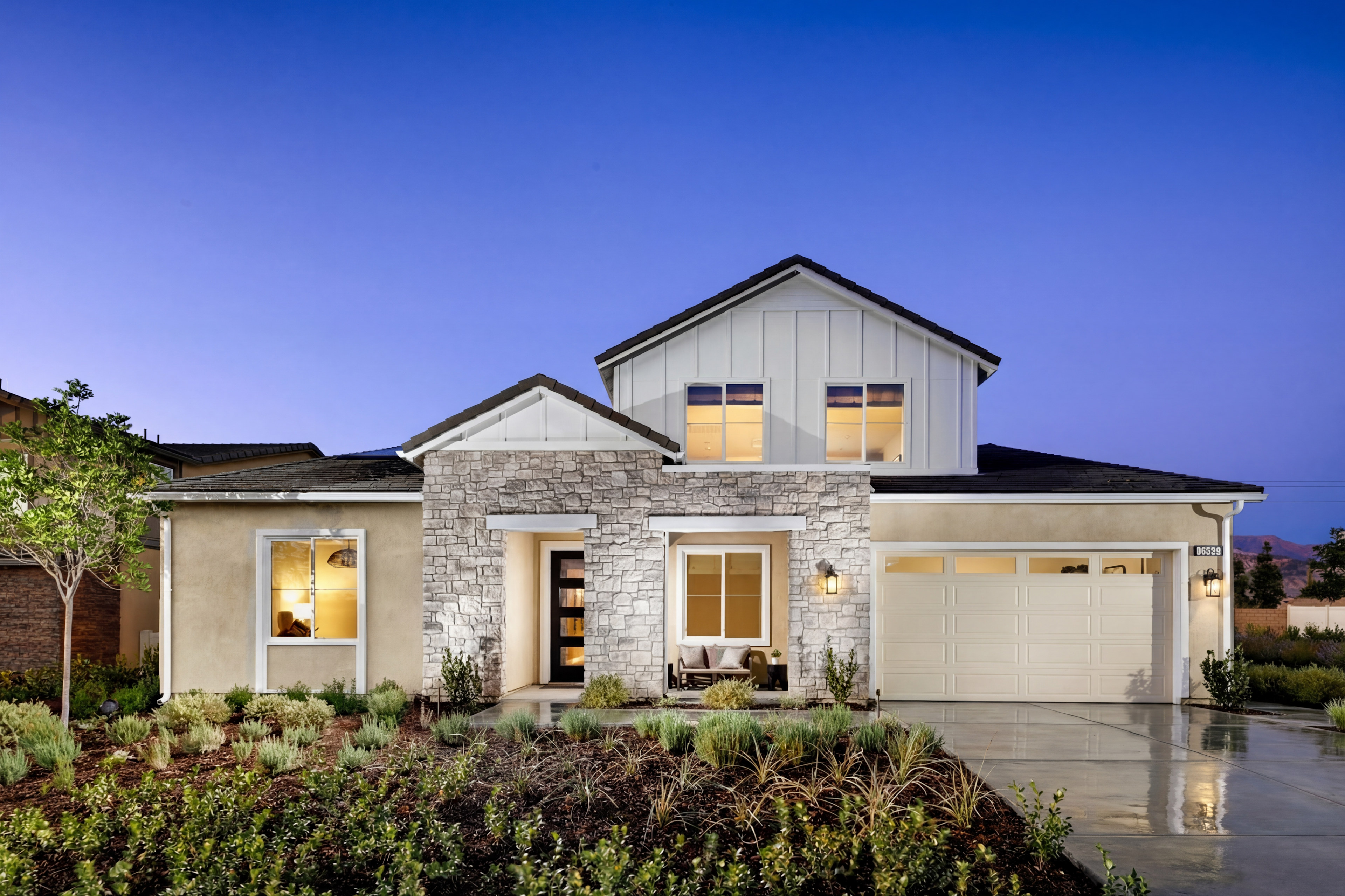 A modern, two-story house with a stone exterior, surrounded by lush landscaping and a paved driveway, set against a clear blue sky.