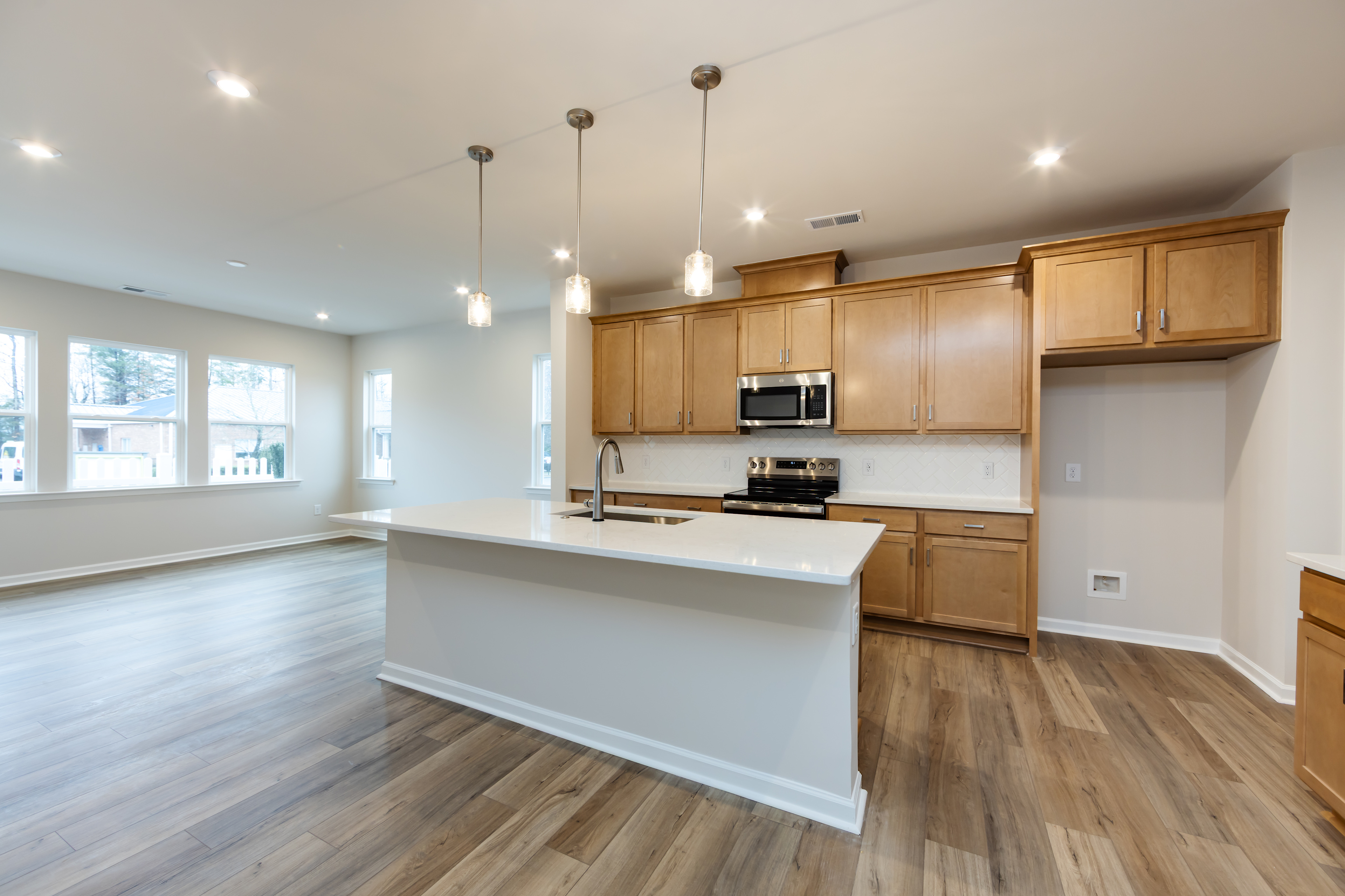 A modern, open-concept kitchen with light wood cabinets, a white island, and hardwood flooring, illuminated by pendant lights.