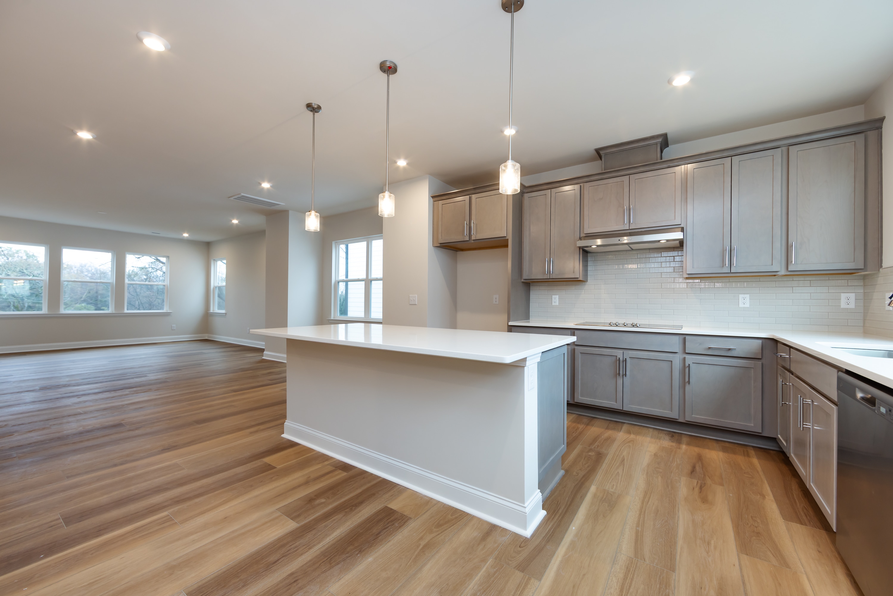 A modern, open-concept kitchen with light-colored cabinets, a white countertop, and hardwood floors, surrounded by large windows that allow natural light to fill the space.
