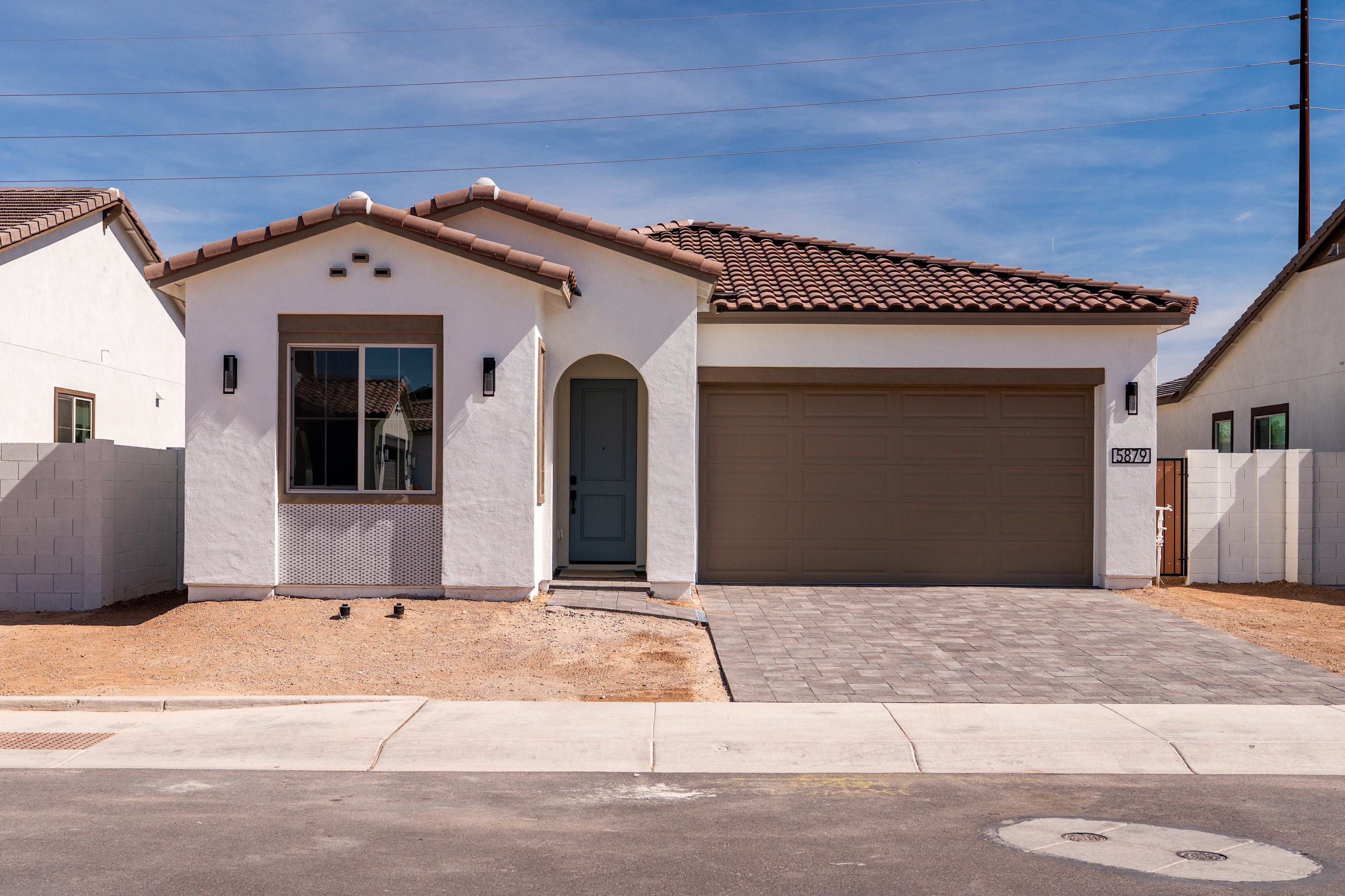 A single-story, Mediterranean-style house with a tiled roof, a garage door, and a paved driveway in the foreground, set against a clear blue sky in the background.