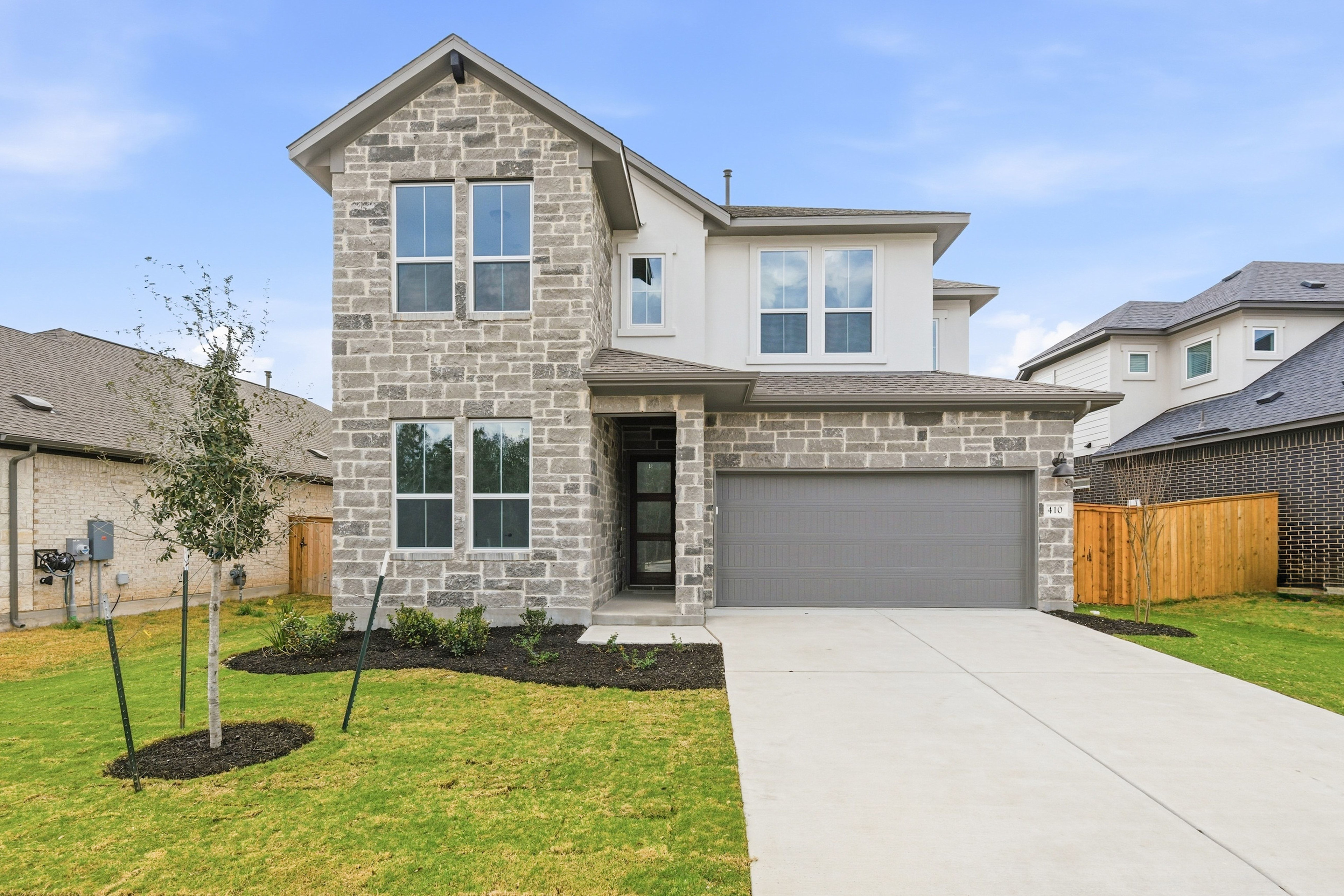 A two-story stone and stucco house with a garage, surrounded by a well-manicured lawn and landscaping, set against a clear blue sky.