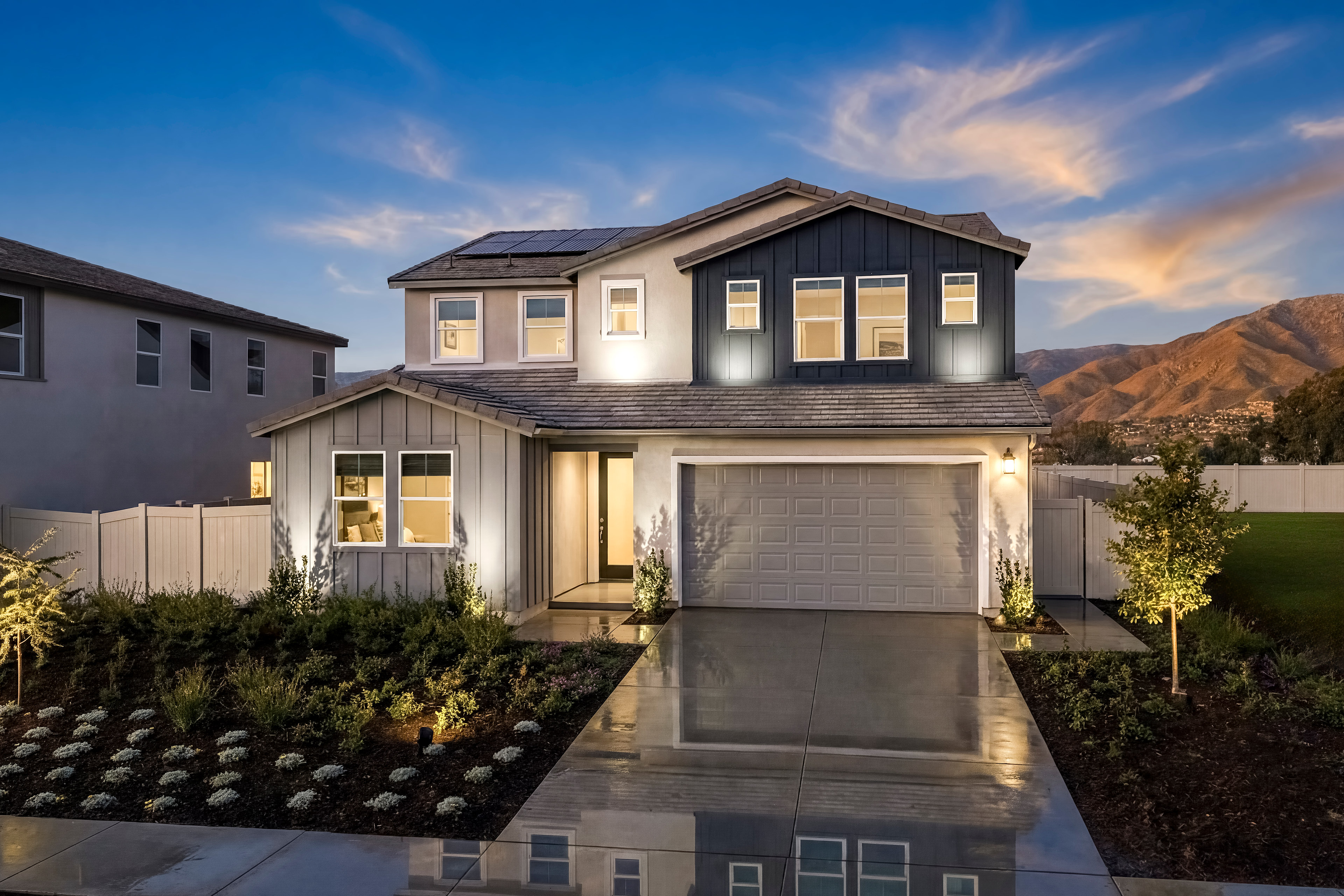 A two-story modern house with a garage, surrounded by landscaped gardens, stands against a backdrop of a vibrant sunset sky.
