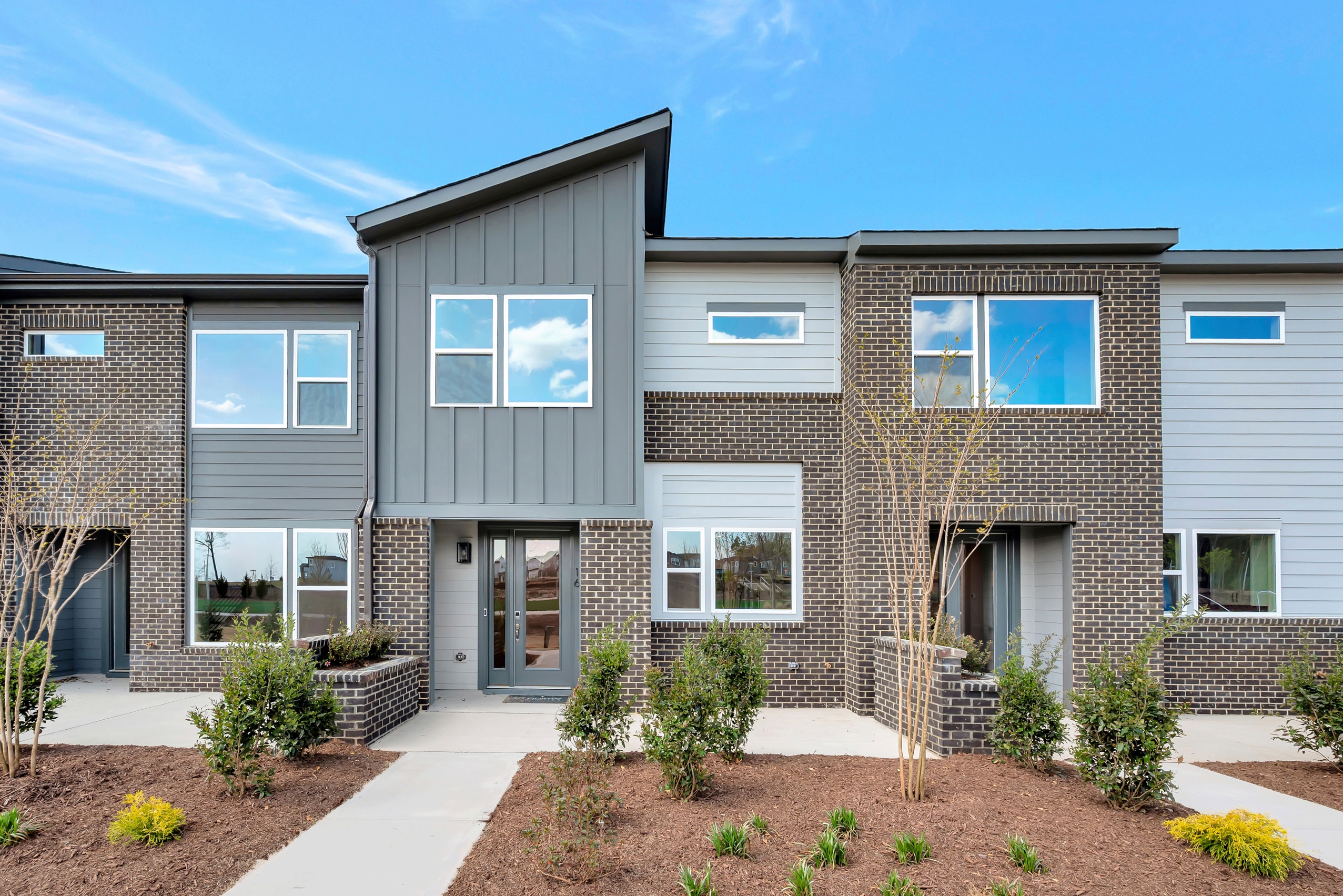 A modern, multi-unit residential building with gray siding, brick accents, and large windows, surrounded by landscaped gardens and a clear blue sky.