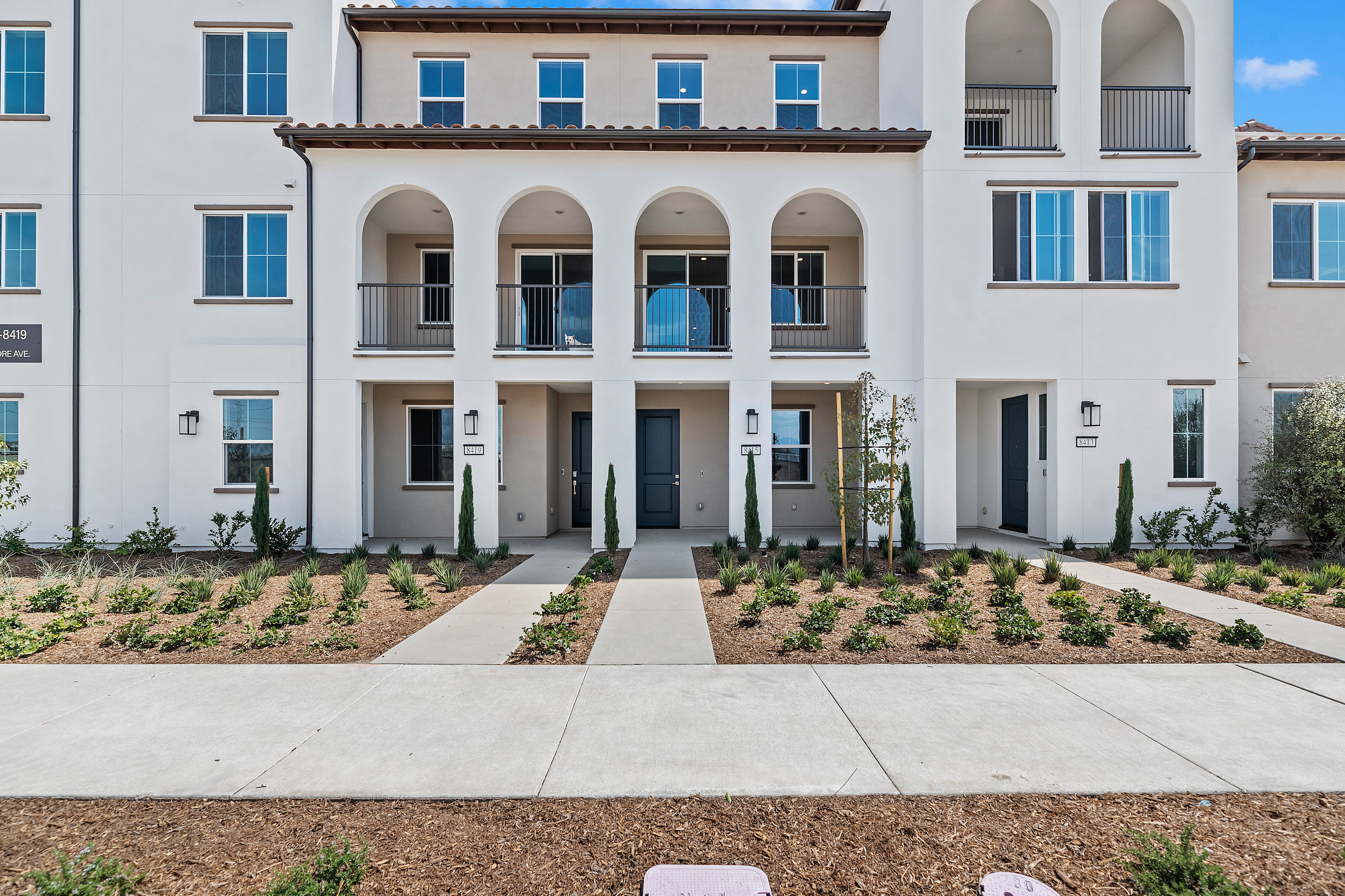 A multi-story residential building with a Mediterranean-style architecture, featuring arched entryways, balconies, and a landscaped front yard with gravel pathways.