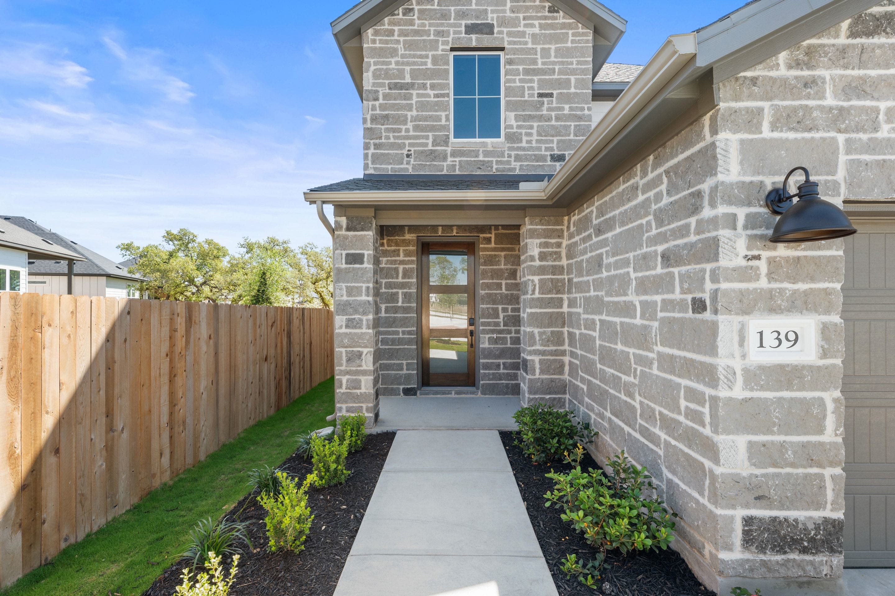 A modern, stone-clad house with a wooden fence and a well-maintained landscaped path leading to the front entrance, set against a backdrop of lush greenery and a clear blue sky.