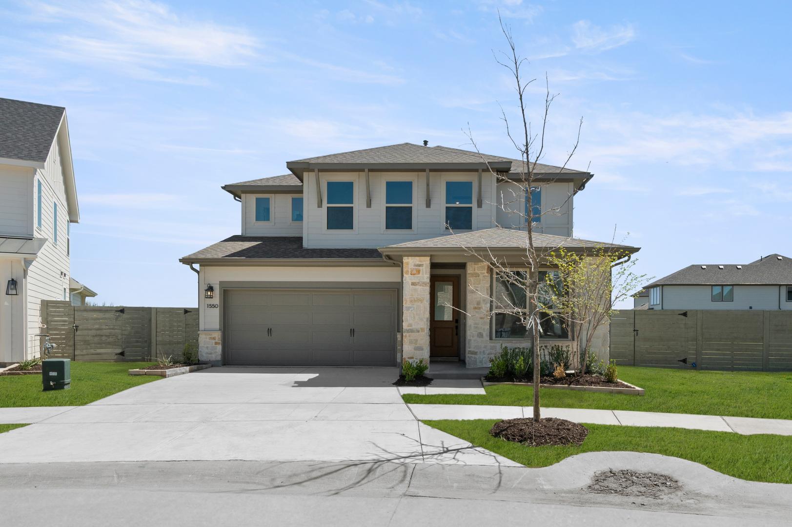 A two-story residential house with a garage, surrounded by a grassy lawn and a paved driveway, set against a clear blue sky with scattered clouds.