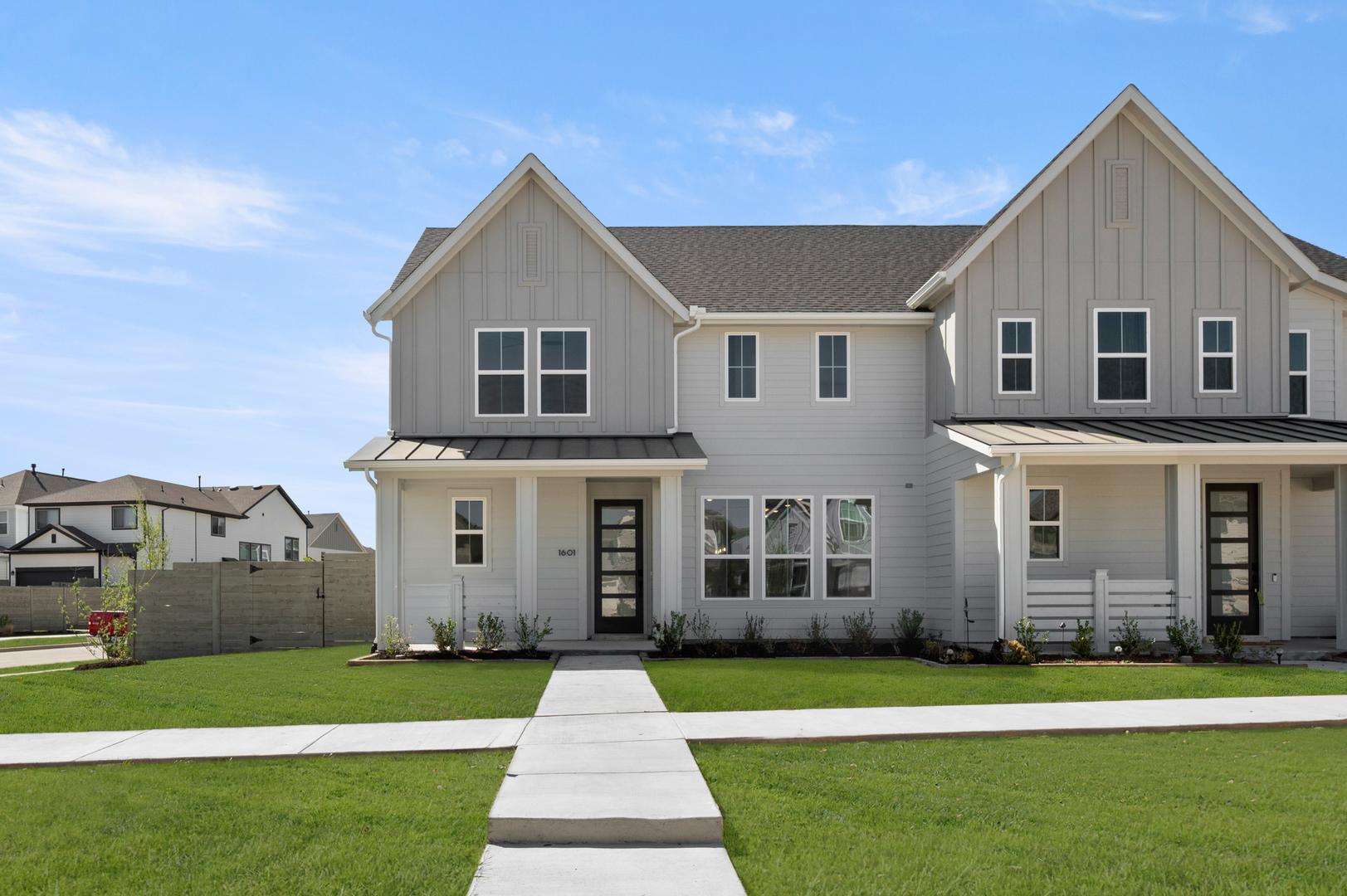 A modern, two-story townhouse with a gray exterior, a well-manicured lawn, and a paved walkway leading to the front entrance, set against a clear blue sky with scattered clouds.