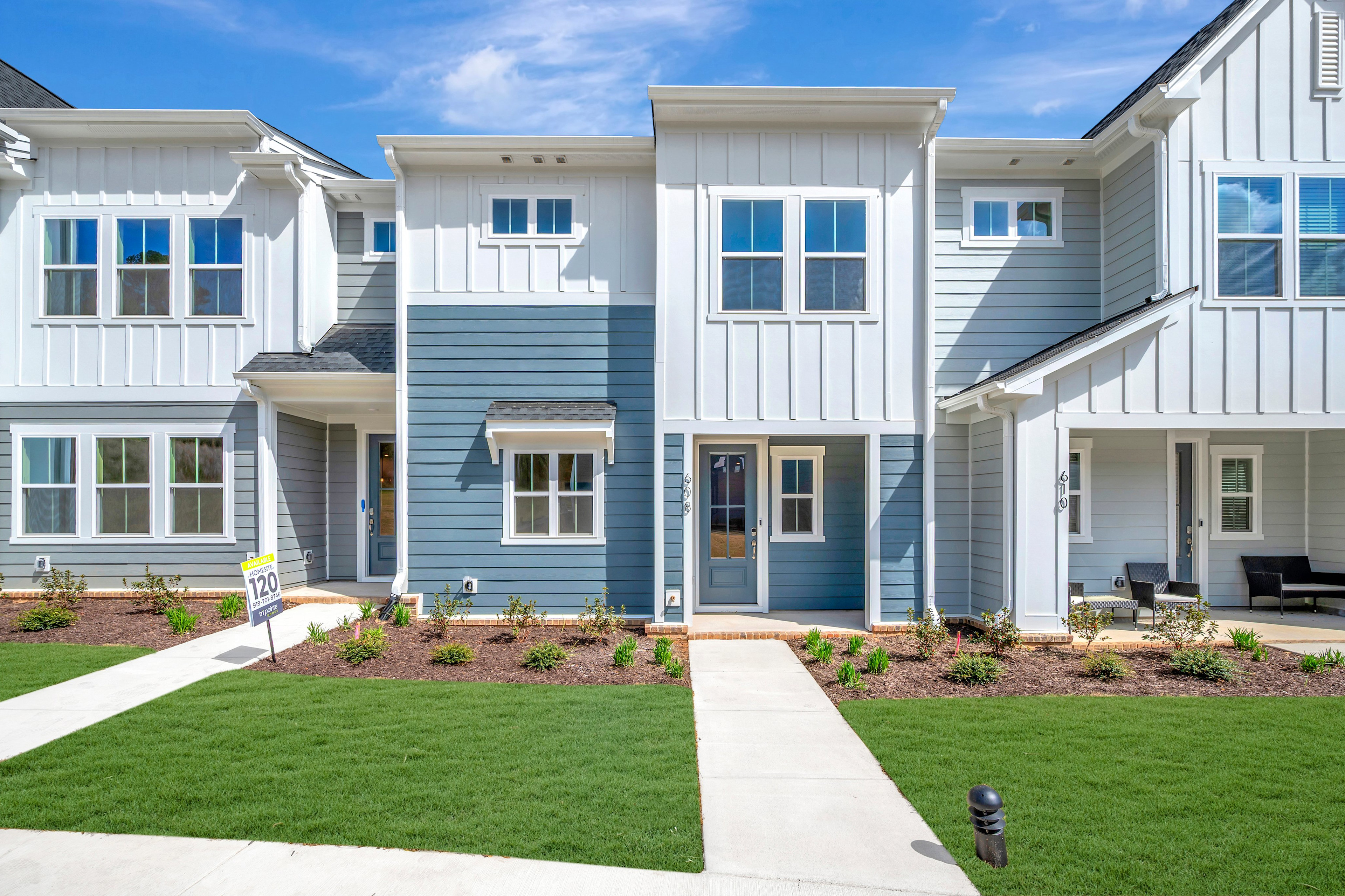 The image depicts a row of modern, two-story townhouses with white siding, blue accents, and well-manicured lawns in the foreground. The sky in the background is a clear, bright blue.
