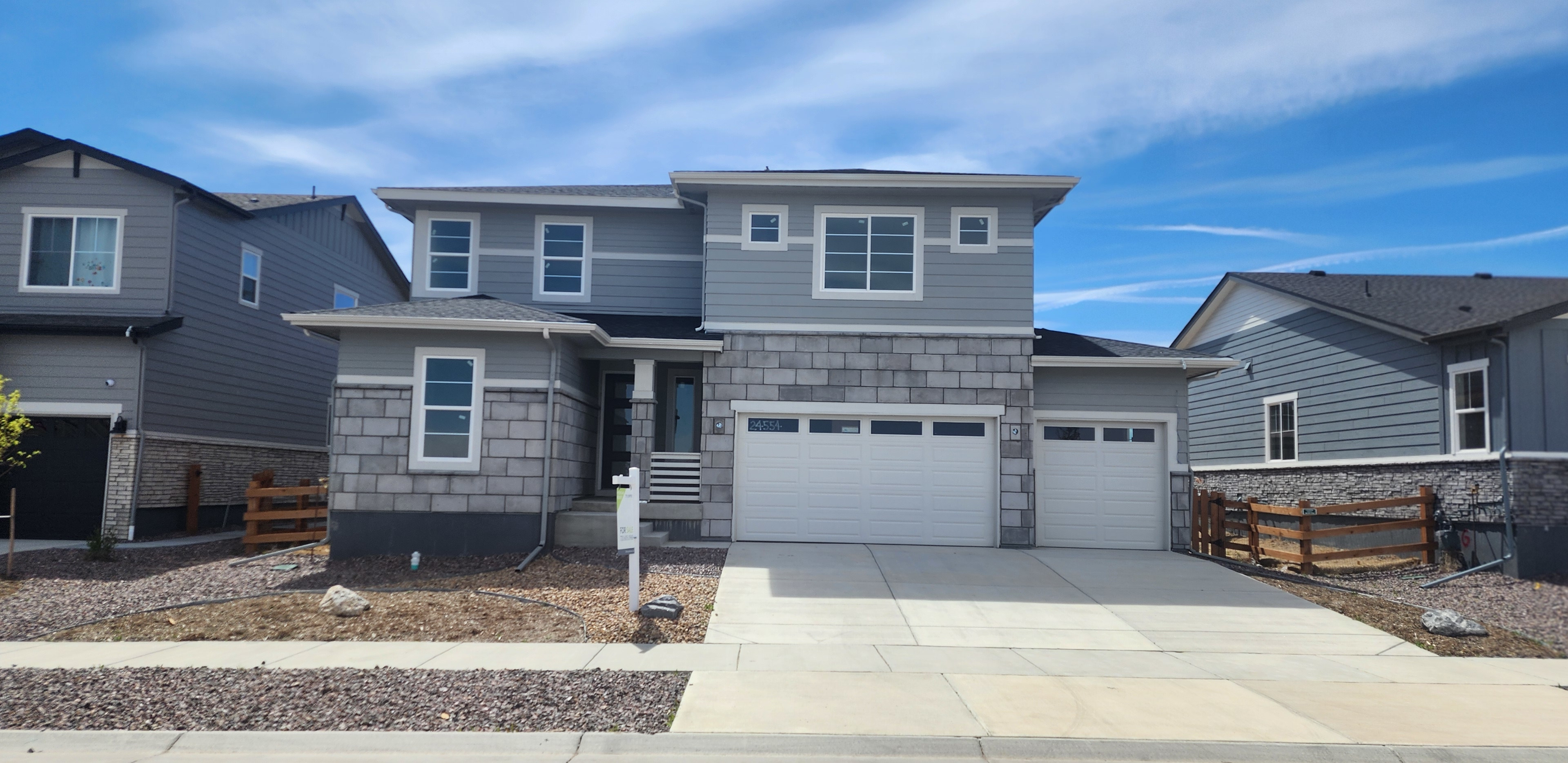 A modern two-story residential house with a gray exterior, a garage, and a paved driveway, set against a blue sky with scattered clouds.