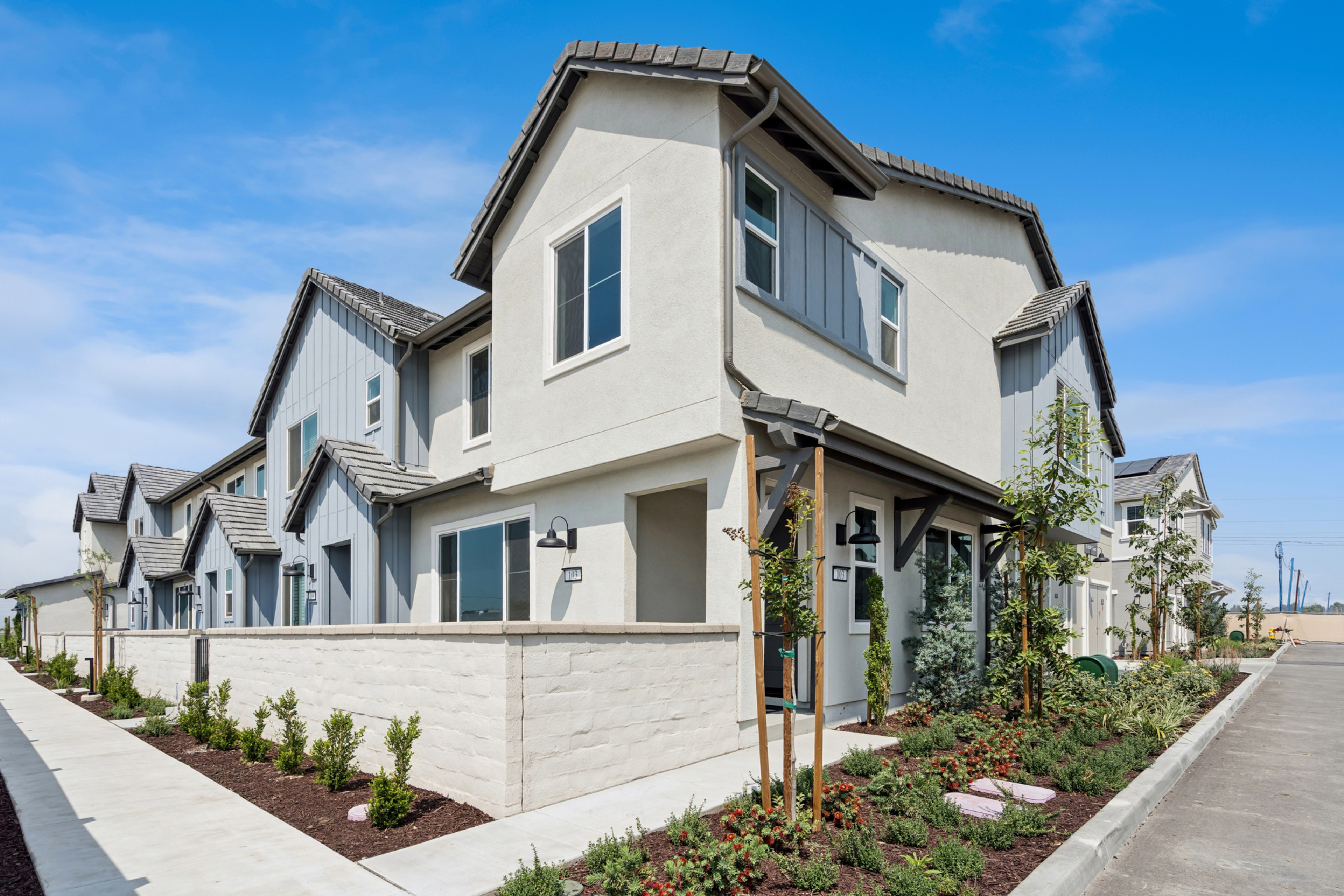 A row of modern, two-story townhouses with gray and white exteriors, surrounded by landscaped gardens and a clear blue sky in the background.