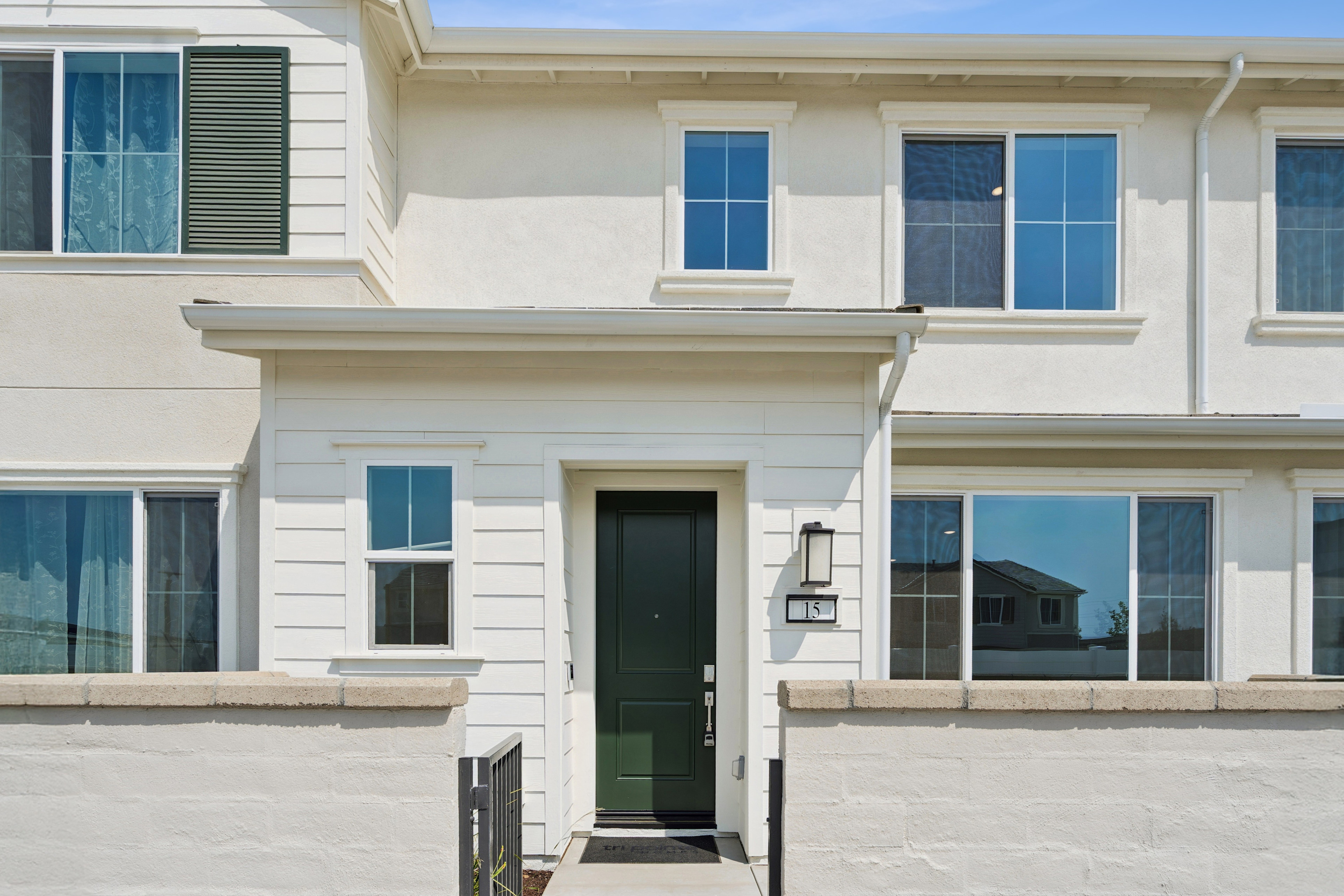 A two-story residential building with a white exterior, large windows, and a front door leading to a paved walkway surrounded by a grassy area.
