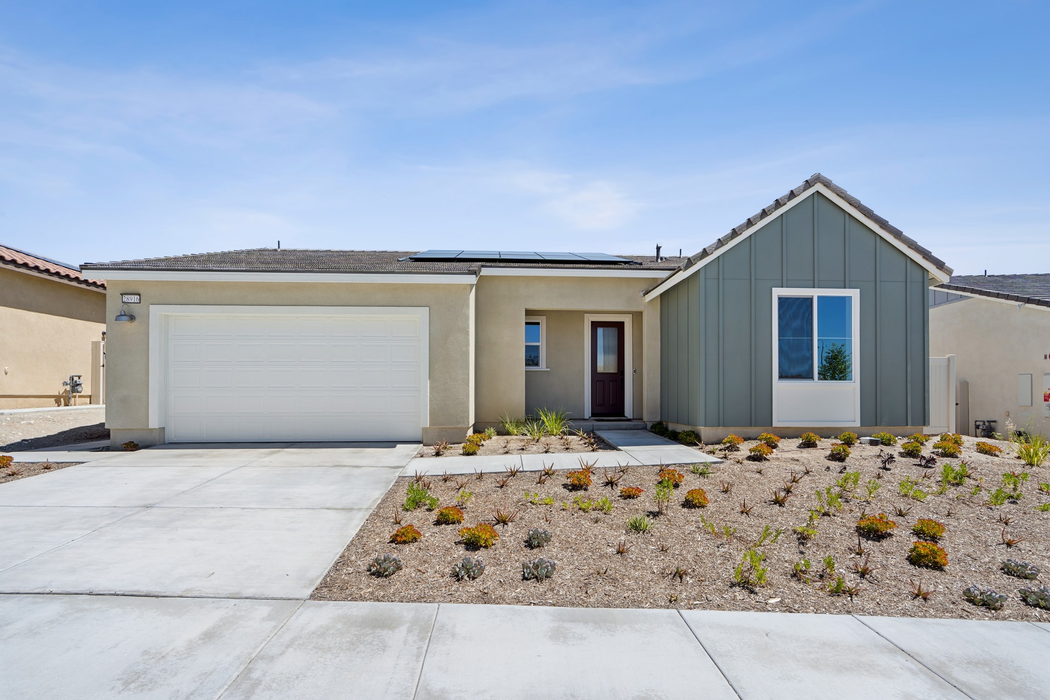 A modern, single-story house with a gray exterior, a white garage door, and a landscaped front yard featuring various plants and a concrete walkway leading to the entrance.