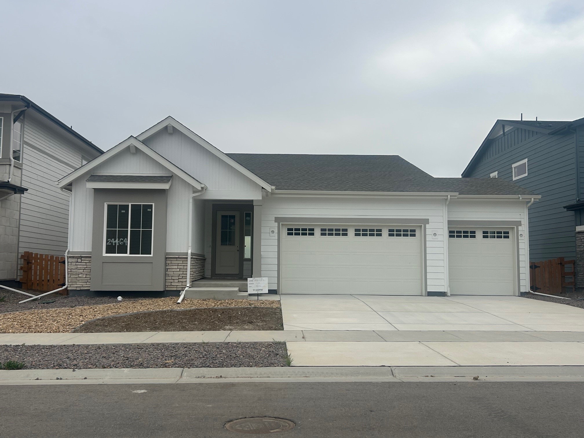 A single-story residential house with a white exterior, a gabled roof, and a three-car garage stands in the foreground, surrounded by a gravel driveway and a paved sidewalk leading to the front entrance.