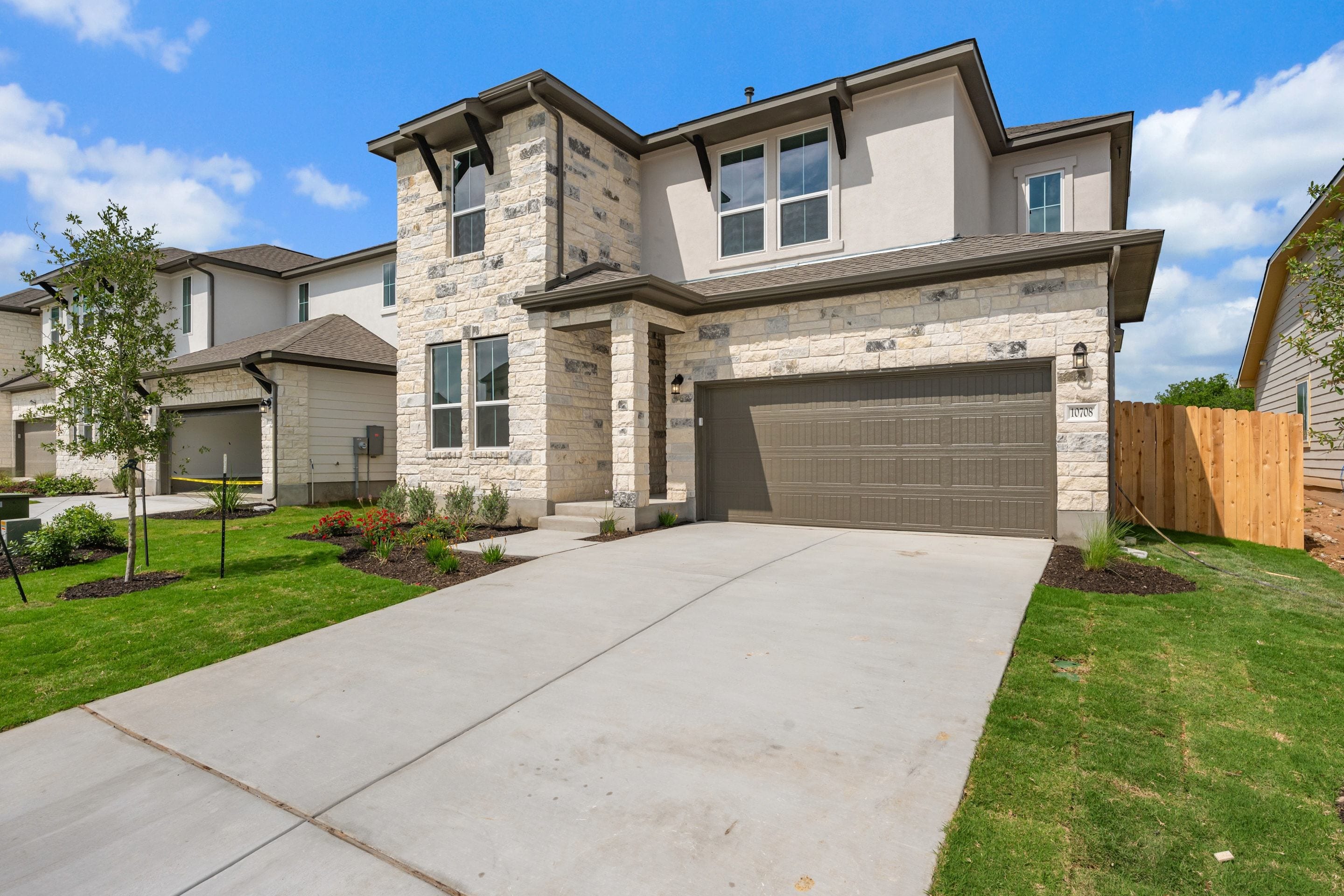 A modern two-story house with a stone exterior, a garage, and a well-maintained lawn in the foreground, set against a clear blue sky with some clouds.