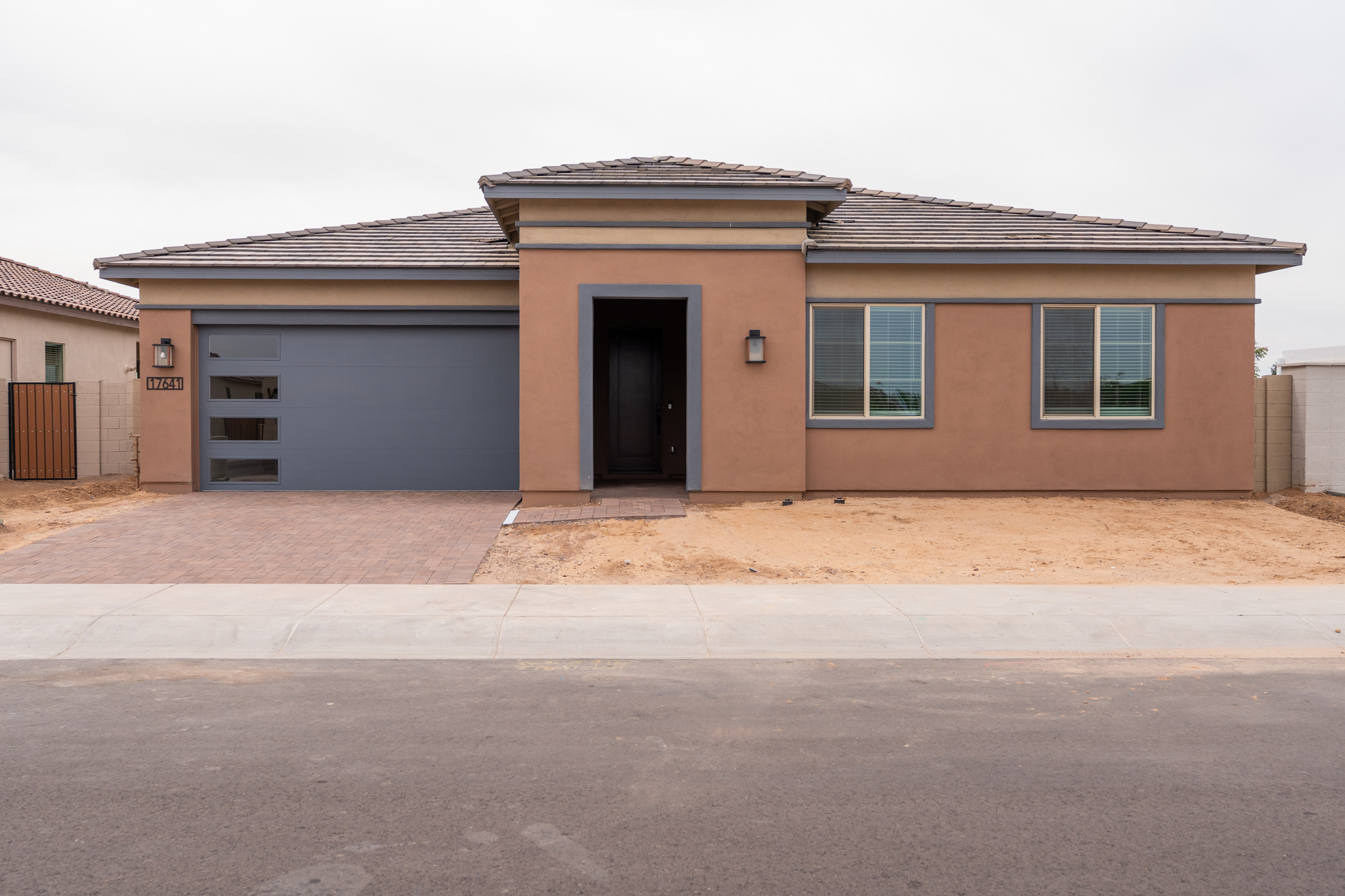A single-story residential house with a tiled roof, a garage door, and several windows, situated on a dirt road with a paved street in the background.